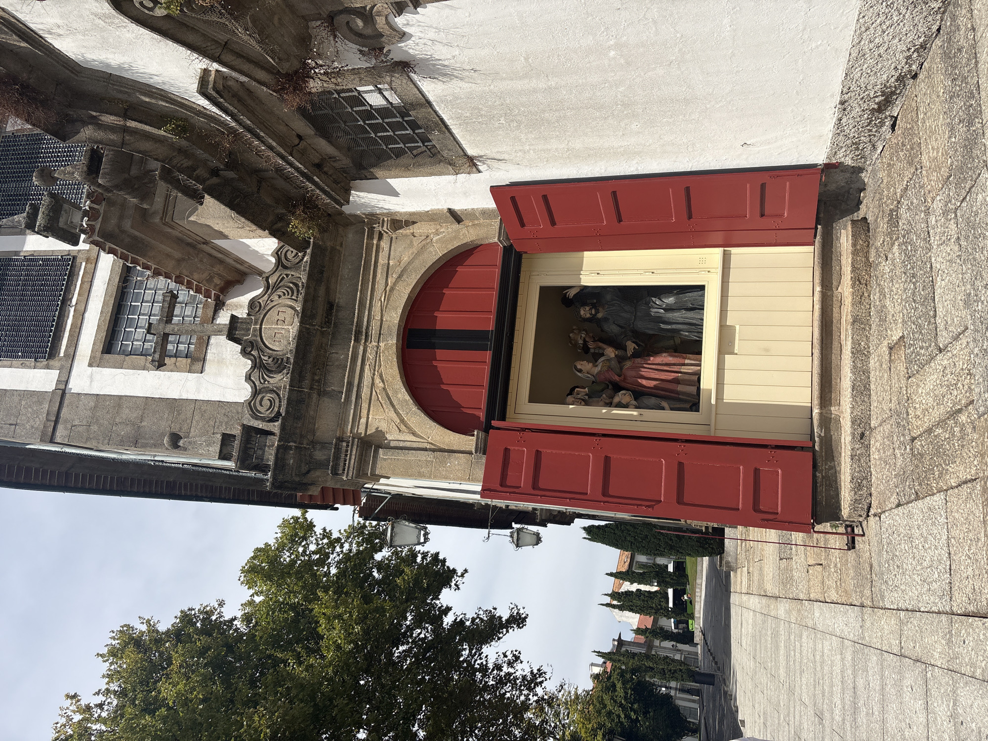 This is a medium-angle outdoor shot of the entrance to a historical building in Guimarães, Portugal, possibly a church or chapel, given the religious statuary visible through the open doorway. The building's facade is white with stone accents and architectural details, including a prominent arched doorway with red shutters. Above the doorway, a stone carving displays the year "1727" and a cross. Inside the doorway, a tableau of religious figures, possibly a scene from the Bible, is displayed. The foreground features a stone sidewalk and steps leading up to the entrance. To the left, a large tree shades a portion of the scene, and beyond that, a street with more trees and buildings can be seen. The sky is overcast. The overall atmosphere is one of historical significance and quiet reverence.