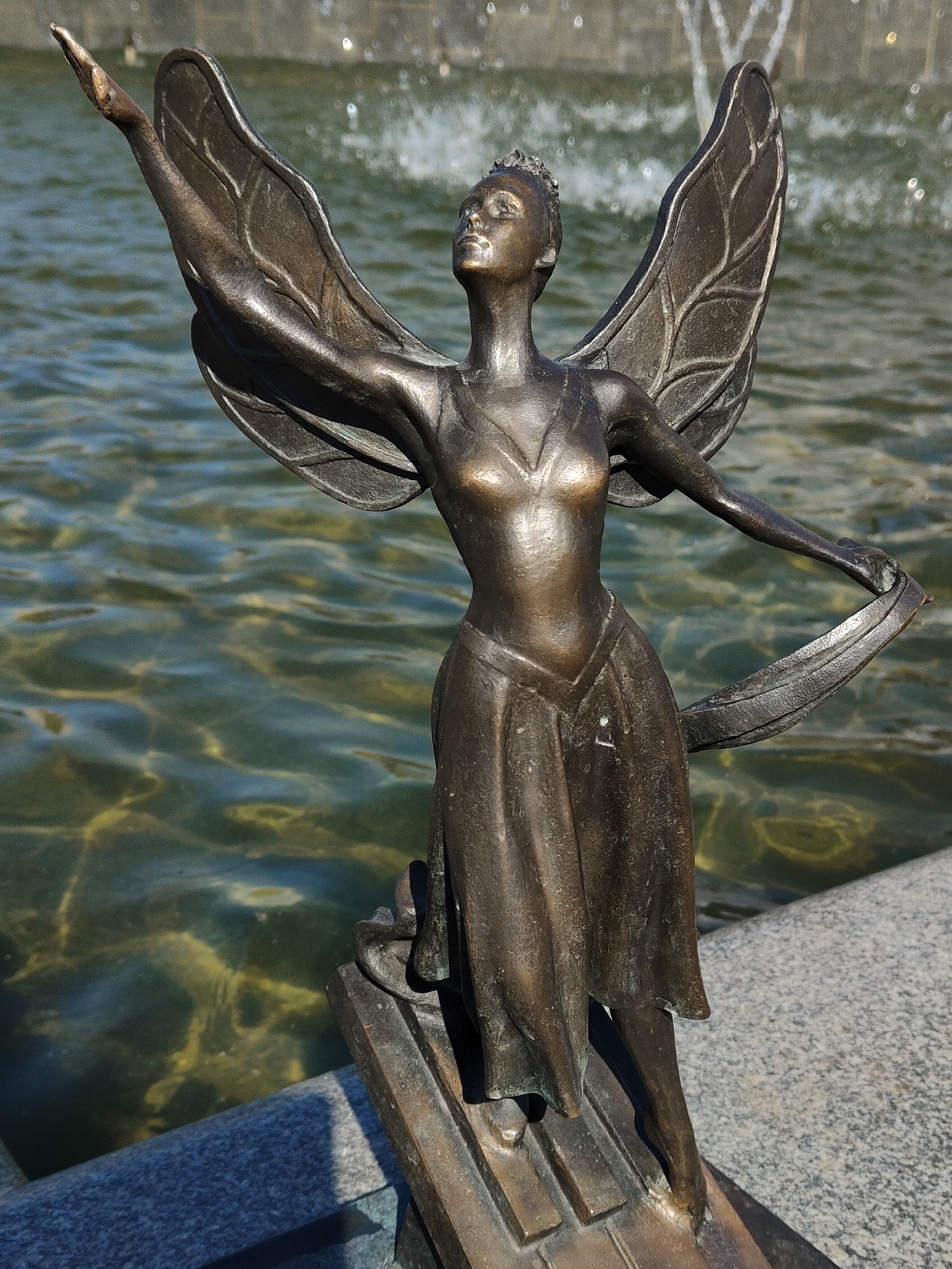 A bronze statue of a winged female figure stands at the edge of a water feature, possibly a fountain, in an unknown city. The figure is depicted with one arm raised and the other extended sideways, holding a flowing piece of fabric or ribbon. She appears to be wearing a dress and a crown or headpiece. The water in the fountain is rippled, and the statue stands on a stone base.