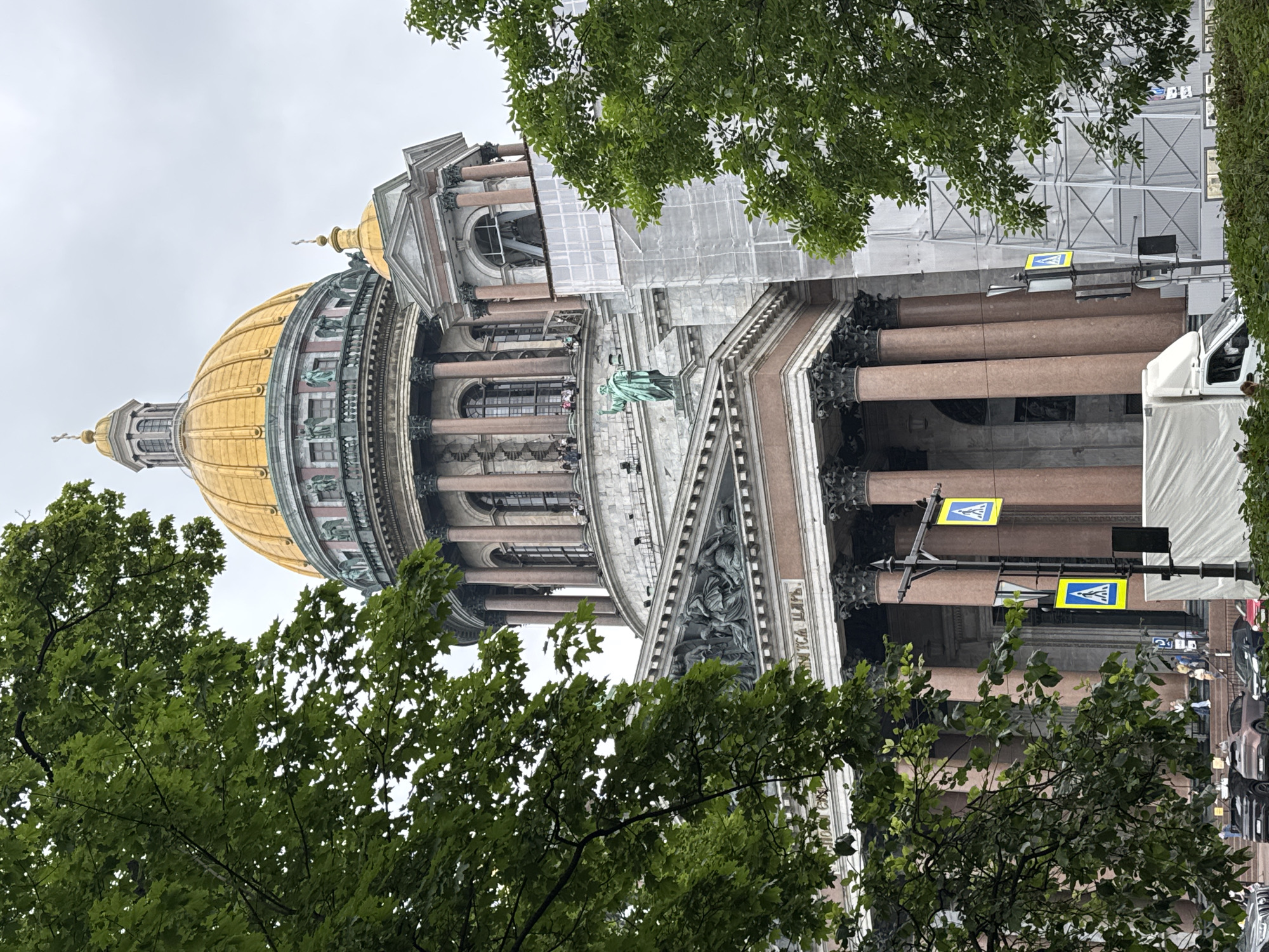 This image shows a side view of Saint Isaac's Cathedral in Saint Petersburg, Russia, partially obscured by green foliage. The cathedral's large, golden dome is visible in the upper portion of the frame, along with smaller domes and architectural details. The building features classical columns and elaborate statues. Scaffolding covered with white sheeting is present on the right side of the building, indicating ongoing construction or renovation. In the foreground, several cars are parked on a street, and pedestrians are visible near the entrance of the cathedral. Two yellow pedestrian crossing signs with blue icons are mounted on a lamppost in the middle ground. The sky is overcast.