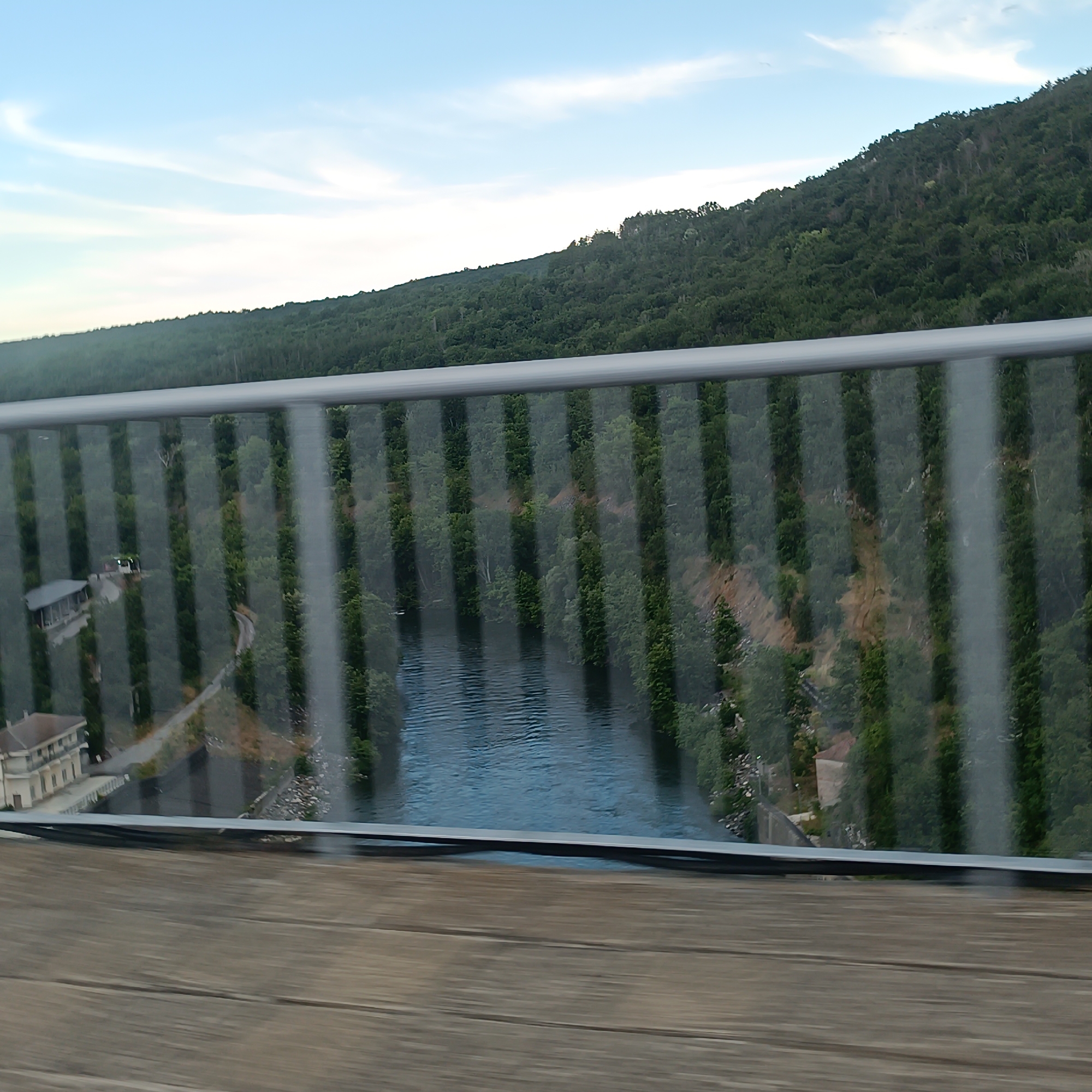 This is a high-angle, medium shot of a river valley with a forested hill in the background. The sky is blue with scattered clouds. A grey metal railing with vertical glass panels runs across the foreground, partially obscuring the view of the scene below. Through the glass panels, a river is visible, flowing through the valley. On the left side of the valley, a building with a light-colored facade and a dark roof can be seen, along with some paved pathways. The hill in the background is densely covered with green trees. The image is taken from a bridge or an elevated viewpoint.