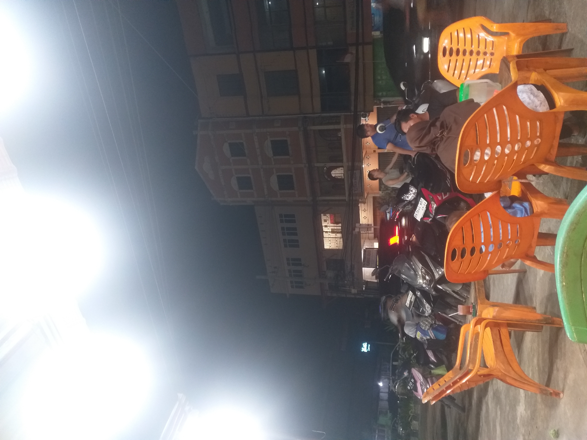 In the city of Dumai, Indonesia, at night, the scene captures an outdoor seating area with multiple orange plastic chairs and tables. A beverage with ice and a straw rests on one of the tables. Behind the seating area, several motorcycles are parked. In the background, a row of multi-story buildings with windows is visible. The lighting in the scene is dim, with a bright light source to the left, creating a hazy effect in that area. Two men are present on motorcycles, one wearing a blue shirt and the other a light-colored shirt. Another person, wearing a dark headscarf, is seated on an orange chair. A sign with a green logo and the word "coffee" in white text is mounted on a pole. The overall atmosphere suggests a casual gathering at a roadside establishment.