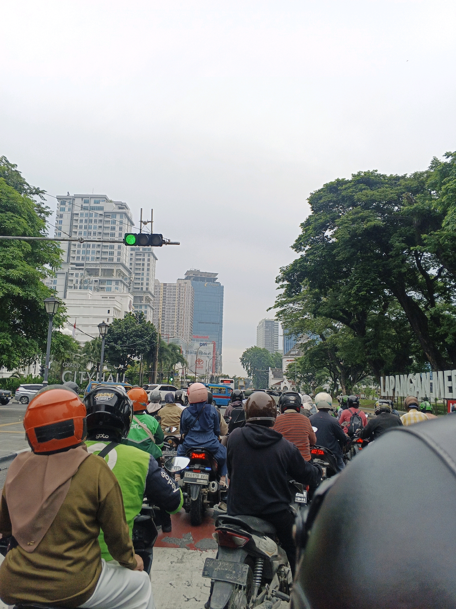 The image depicts a street scene in the City of Medan, Indonesia, characterized by a traffic jam of motorcycles. The foreground is dominated by the backs of several motorcyclists wearing helmets of various colors including orange, black, and brown. A significant number of motorcycles are queued up, suggesting a standstill or slow-moving traffic. In the mid-ground, more motorcycles and a blue bus are visible. Tall buildings, including a modern skyscraper with many windows and another with a blue glass facade, rise in the background, indicating an urban setting. A prominent traffic light displays a green signal. Large trees with dense foliage line the right side of the street, providing a stark contrast to the urban architecture. A sign is partially visible on the right side with text that appears to read "LARANGAN SAN MENER". The sky is overcast and bright. The overall atmosphere is one of congested urban traffic on a cloudy day.