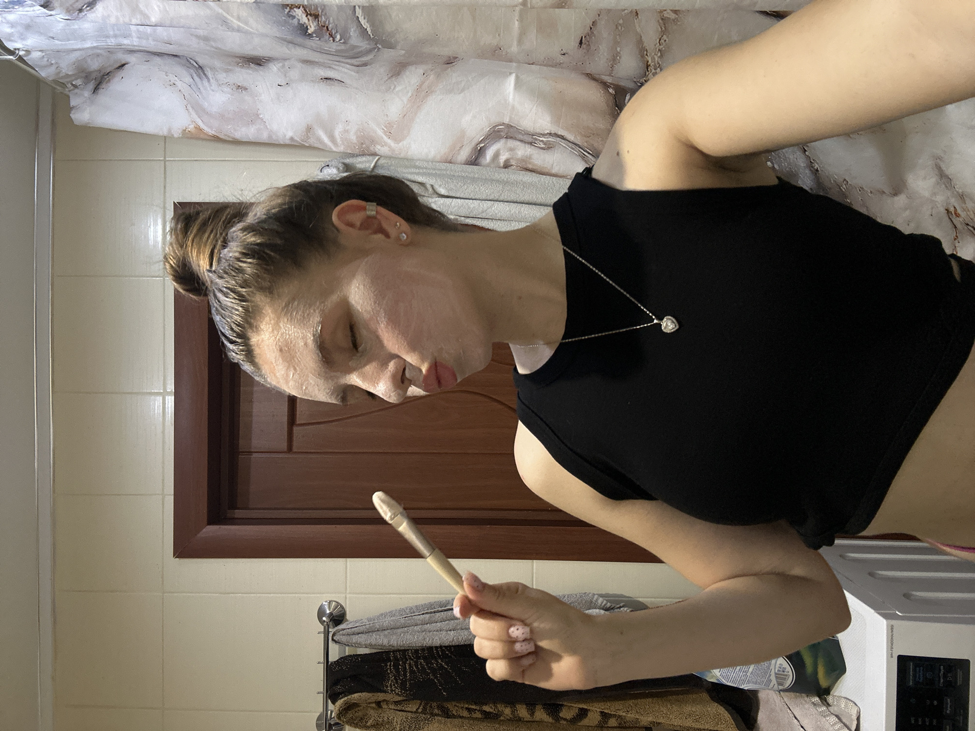 A woman is depicted in a bathroom setting, likely in a residential building. She is holding a cosmetic brush, with what appears to be a facial mask applied to her face. She is wearing a black tank top and a necklace. The bathroom has tiled walls, a brown door or window frame, and a shower curtain with a marble-like pattern. A towel rack with towels is visible. A washing machine or dryer is present in the lower right corner of the image. There is no immediate information in the image to confirm the city of Klin, Russia.