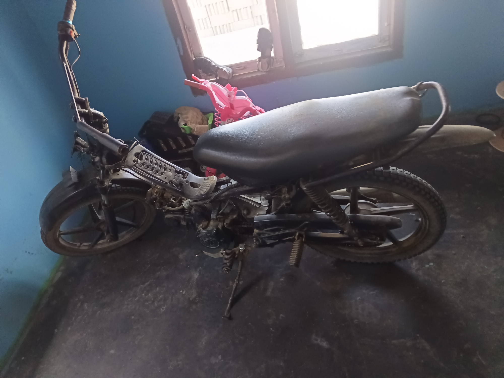 The image shows a black motorcycle parked indoors with a blue wall and a window in the background. The motorcycle is a dirt bike or scooter style with a visible kickstand and exhaust pipe. A pink child's seat is attached to the rear of the motorcycle. The floor is a dark, rough surface, possibly concrete. There are no people visible in the image. The scene appears to be a storage area or a workshop.