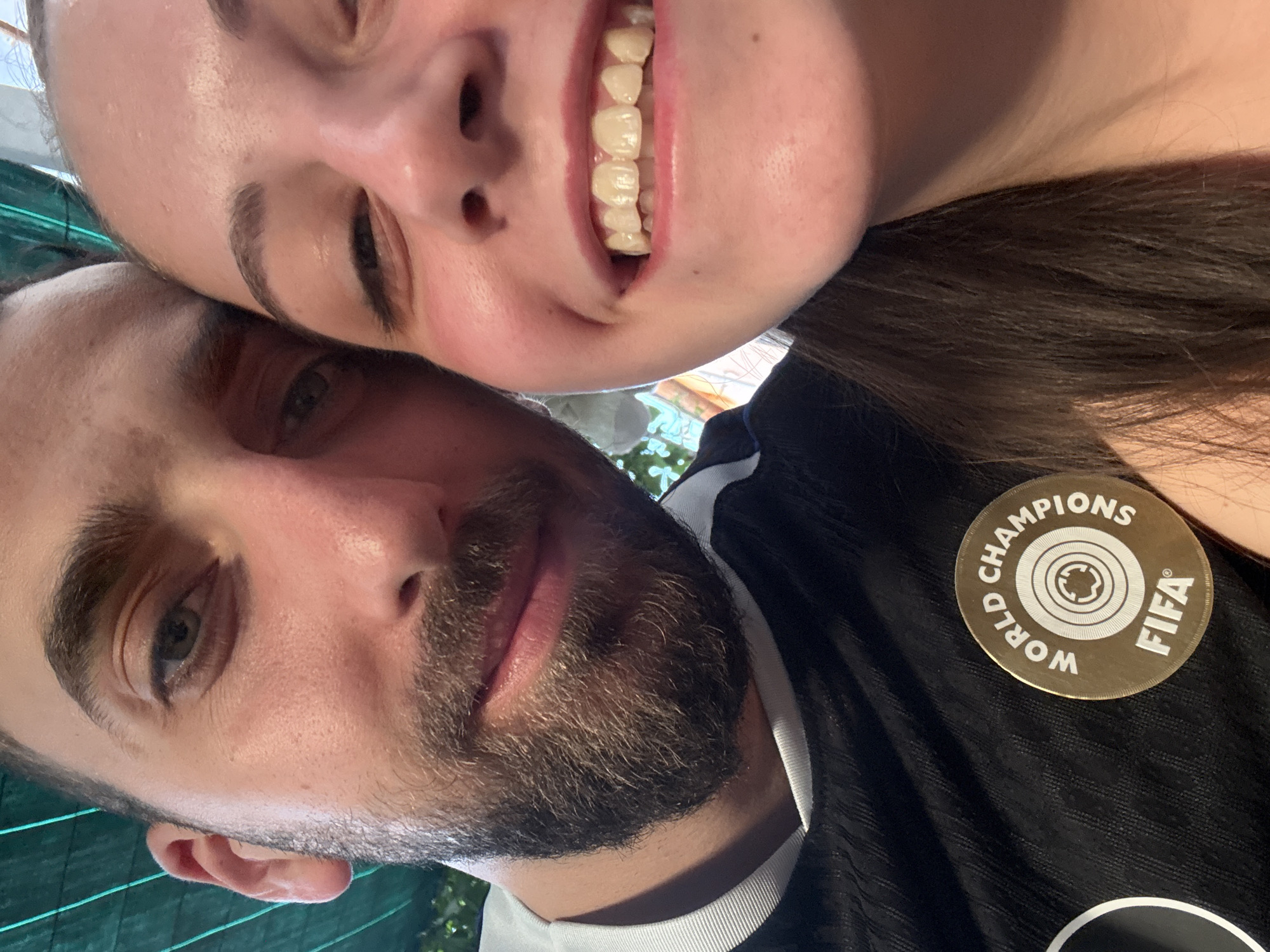 A selfie-style photograph features a man and a woman posing closely together. The woman is above and slightly behind the man, smiling broadly with her head tilted. The man, with a beard, is positioned lower in the frame, looking up towards the camera with a more neutral expression. He is wearing a black sports shirt with white trim and a circular gold FIFA World Champions patch. The background is obscured, though hints of greenery suggest an outdoor setting. The location cannot be identified as Tsarevo, Bulgaria from the visual data provided.
