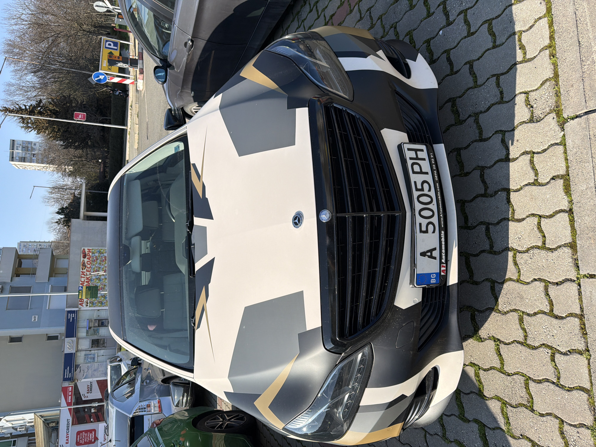 A Mercedes-Benz E-Class sedan (W212 facelift), featuring a custom geometric camouflage vinyl wrap in white, light grey, dark grey/black, and gold, is parked head-on in a paved lot in Burgas, Bulgaria. Its front license plate displays "BG A 5005 PH". The vehicle's interior appears empty.
The parking surface consists of interlocking light grey concrete pavers with moss growing in the gaps, illuminated by direct sunlight which casts distinct shadows. To the left of the Mercedes is a partially visible green car, followed by a silver Toyota Prius. To the immediate right is a dark grey sedan.
In the background, a light blue multi-story residential building with commercial ground-floor units, including "EASYCREDIT" signage, is visible to the left. To the right, a low wall with a billboard advertising "АВТОЧАСТИ АКСЕСОАРИ" (Auto Parts Accessories) separates the lot from a street. Across the street, bare deciduous trees, streetlights, a yellow "BILLА" parking payment machine, and various road signs (including a blue directional arrow and a 'P' for parking sign) are present. A tall, slender residential tower stands in the distant background. The sky is clear and blue. No individuals are visible in the scene.