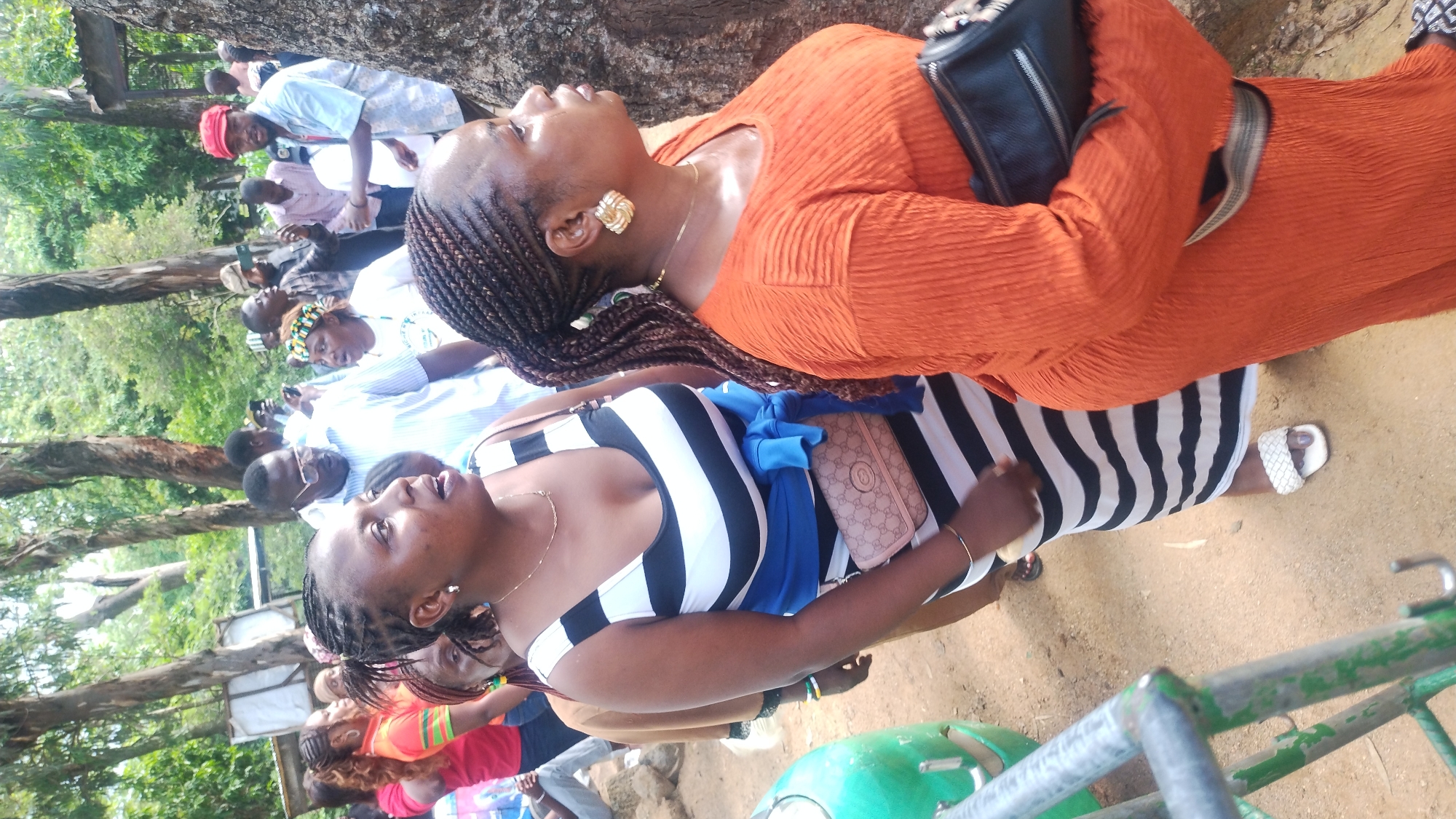 In Jos, Nigeria, a group of people are gathered outdoors in a natural setting with trees and dirt ground. The focus is on two women in the foreground. The woman on the left wears a black and white striped dress with a blue sash tied around her waist, and carries a beige purse. The woman on the right is dressed in an orange, textured dress and holds a black clutch purse. Other individuals, including men and women, are visible in the background, some holding cameras and phones. The overall scene suggests a social gathering or event taking place in an outdoor urban or semi-urban environment.