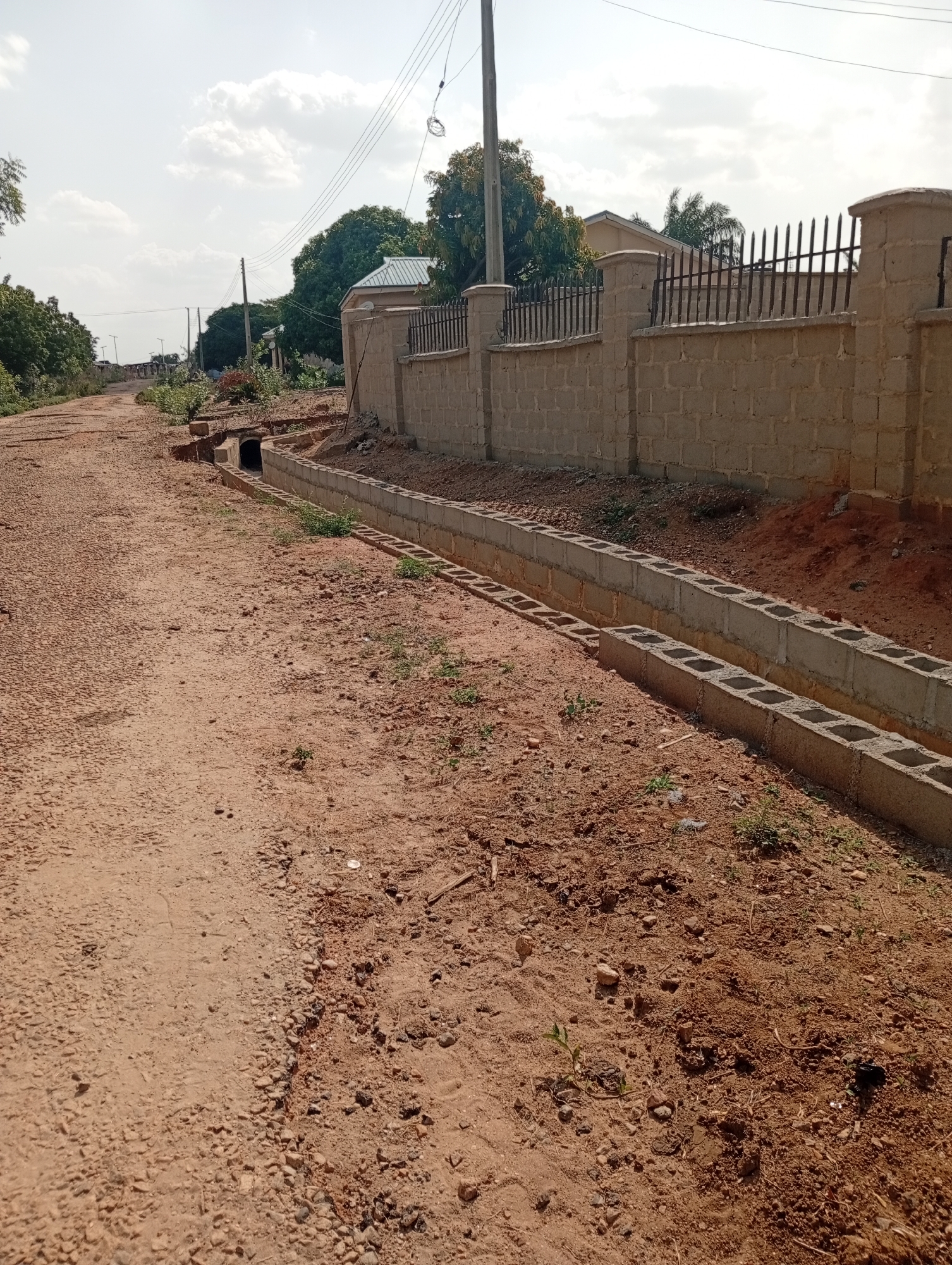 The image depicts a dirt road in Adankolo, Nigeria, bordered by a concrete block drainage ditch. A partially constructed concrete block wall with a black wrought-iron fence runs along the right side of the road. A drainage culvert is visible on the left side of the road, leading under the road. Power lines are overhead. The sky is overcast. The road surface is dry and dusty.