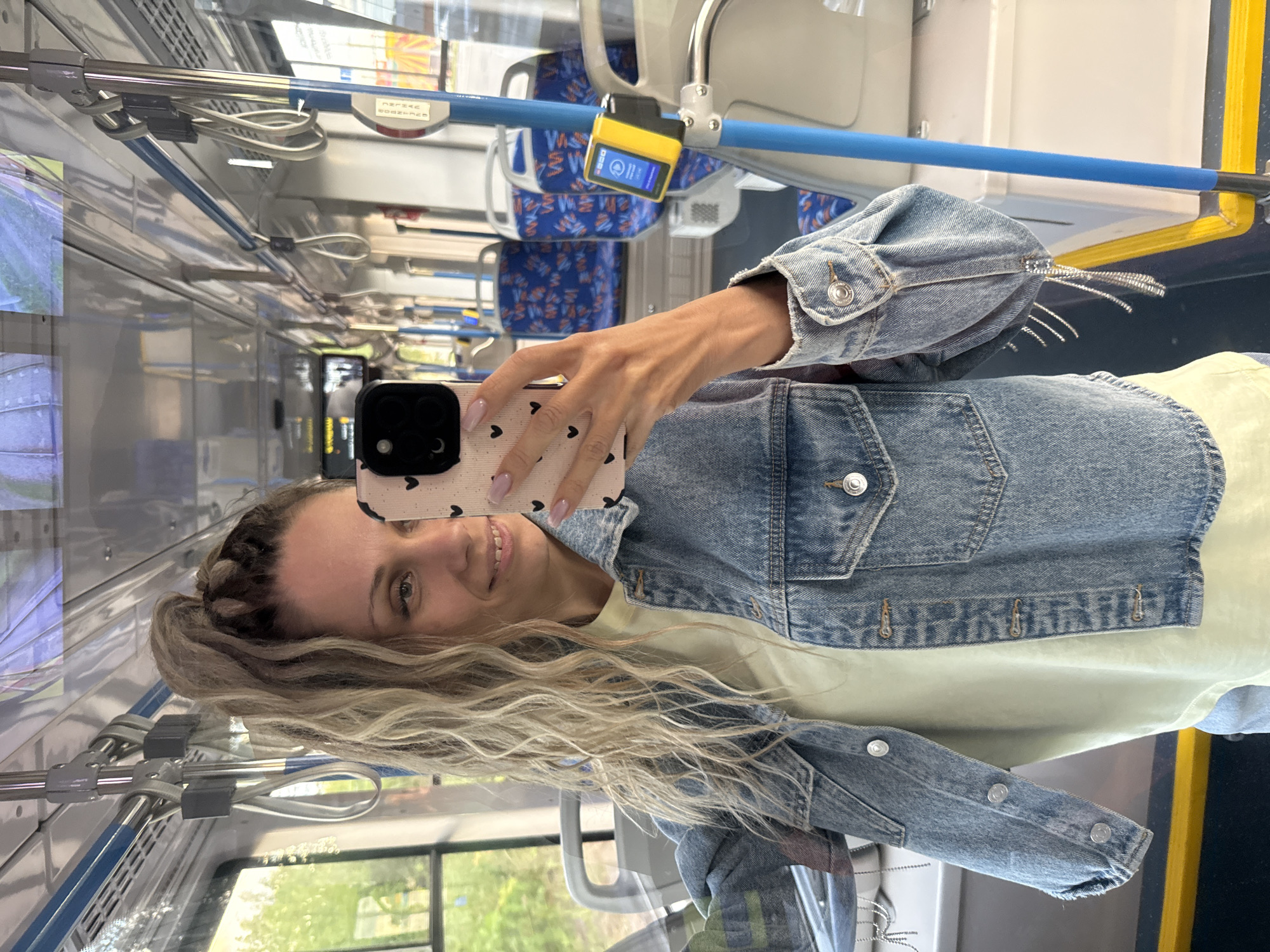 A woman with blonde, curly hair pulled into a high ponytail takes a selfie on a bus in Rusanovo, Russia. She is wearing a denim jacket over a light yellow t-shirt. The bus interior is visible, with blue and white seats and metal grab bars. A yellow ticket validator is mounted on a blue pole.