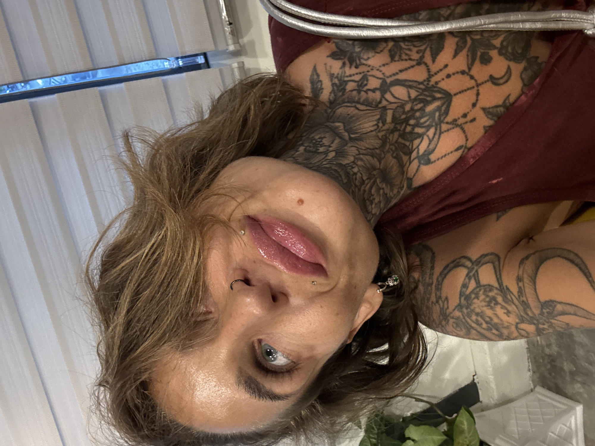 The media file features a close-up, rotated portrait of a woman with light brown, tousled hair. She has several facial piercings, including a nose ring and a lip ring. Her eyes are a striking blue, and she appears to be looking towards the camera. Her lips are slightly parted, and she is wearing a glossy pink lipstick.
The woman is adorned with extensive tattoos on her neck and arm, featuring floral and possibly animal motifs. She is wearing a maroon-colored top, and a metallic silver strap is visible across her chest. The background is out of focus, but appears to be indoors, with vertical blinds on the left and what looks like a white, textured surface on the right. There are also hints of green foliage in the lower right corner.
The lighting suggests it is daytime, possibly indoors with natural light coming from behind the blinds. The overall mood is intimate and personal, highlighting the subject's features and body art. No text is visible in the image.