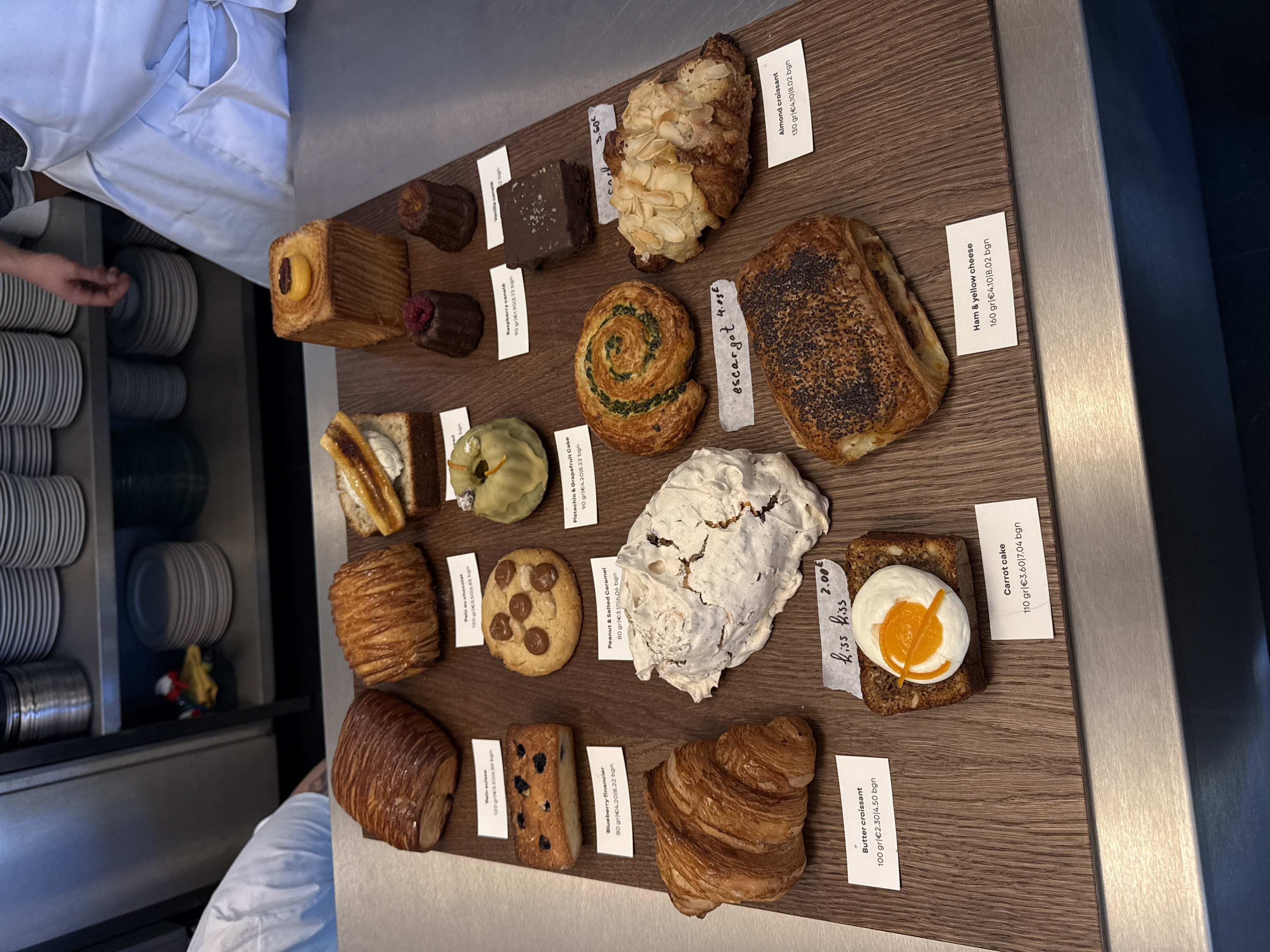 This image captures a close-up, overhead view of a bakery display counter in Sofia, Bulgaria. A professional assortment of approximately sixteen baked goods and pastries is arranged on a textured light brown wooden board. The board rests on a reflective stainless steel counter surface in the immediate foreground.
Each pastry is accompanied by a small white rectangular label providing its name, weight in grams, price in Euros (€), and price in Bulgarian Leva (BGN). From left to right and top to bottom, visible items include: a Pain Suisse, a Pain au chocolat, a slice of banana bread topped with cream and caramelized banana, and a large rectangular bread piece topped with a small yellow custard tartlet. Below these are a blueberry financier, a round peanut and salted caramel cookie, a green-glazed pistachio and grapefruit bundt cake, a raspberry canelé, a vanilla canelé, and a dark chocolate brownie sprinkled with sea salt. Further down, a classic butter croissant, a large white meringue with a cracked surface, a round escargot pastry with a green filling, and an almond croissant are displayed. In the bottom row, a slice of carrot cake with white frosting and an orange curl is visible, accompanied by a handwritten "kiss kiss 2.00€" label, and a rectangular pastry topped with poppy seeds, identified as a ham and yellow cheese pastry.
In the background, two individuals are partially visible. On the far left, a person's forearm and hand, dressed in a white uniform, are positioned near the counter. On the far right, a person's torso and arm, wearing a white apron over a grey long-sleeved shirt, stand behind the counter. Behind these individuals, dark grey shelving units are stacked with numerous white plates and various metal containers, indicating a functional kitchen or service area. A small, multicolored object rests on a yellow cloth on one of the shelves.
