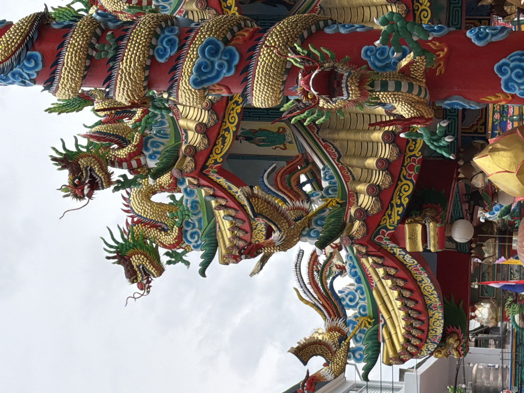 This image captures a detailed close-up of ornate, colorful dragon sculptures adorning the exterior of a building, likely a temple or cultural site, in Chiang Mai City Municipality, Thailand. The dragons are intricately designed with golden scales, red bodies, and vibrant green and blue accents, evoking a sense of power and mystique. Some dragons are depicted with their mouths open, as if roaring, while others are shown coiled or in motion. The sculptures are part of a larger, elaborate architectural decoration, featuring swirling cloud motifs and decorative patterns in yellow and blue.
The lighting suggests it is daytime, with diffused light indicating an overcast sky. The weather appears to be cloudy, which is common in Chiang Mai. The overall scene is rich in cultural symbolism, with dragons being a significant motif in East Asian art and mythology. There is no visible text in the image. The focus is on the artistic and architectural craftsmanship of the dragon sculptures and the building's facade. The perspective is from below, looking upwards at the detailed artwork, emphasizing its grandeur and complexity.