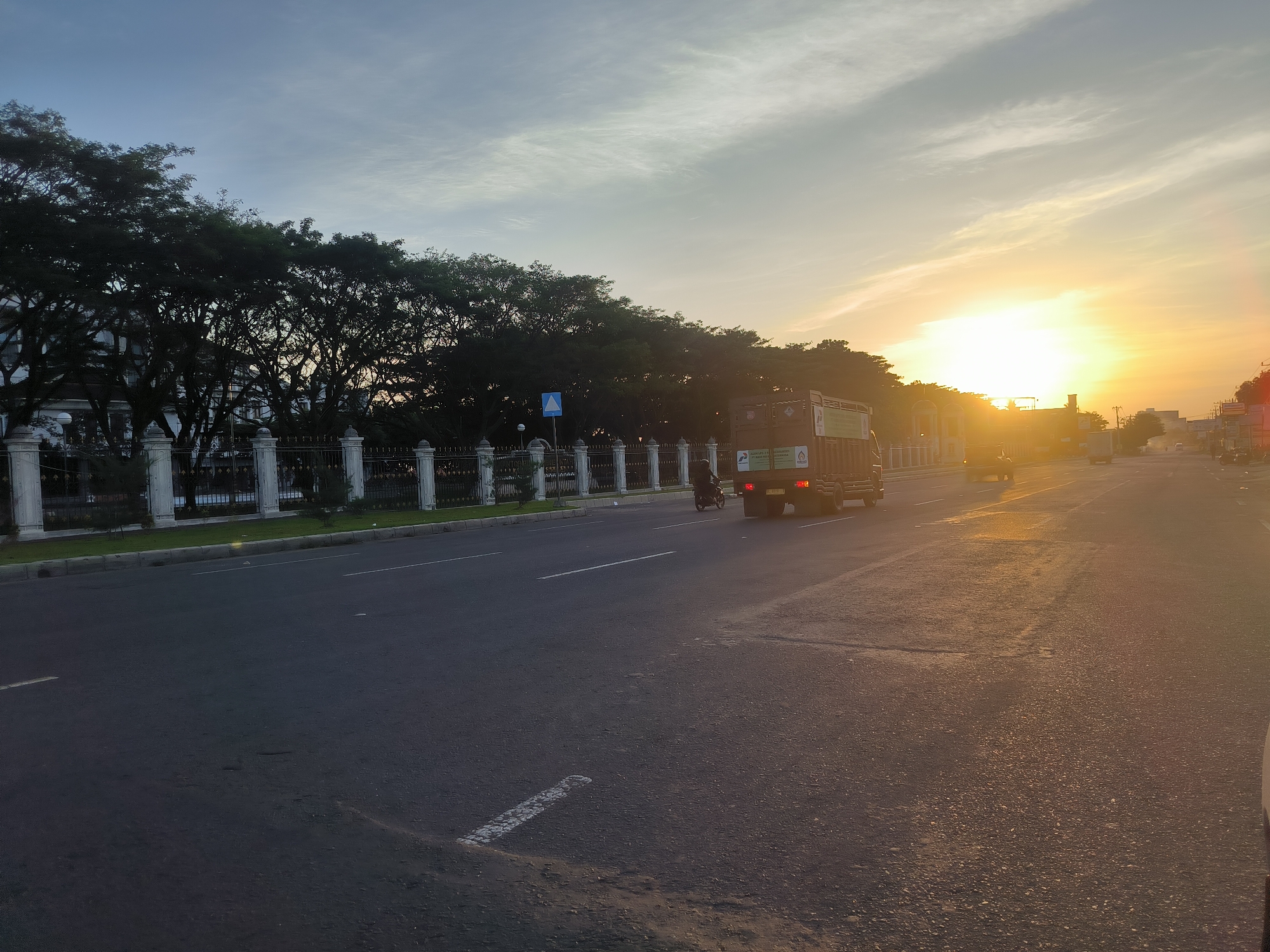 A street in Cotgapu, Indonesia is bathed in the warm light of the setting sun. On the left side of the road is a white building with a decorative fence, lined with trees. A blue pedestrian crossing sign is visible beside the fence. A truck drives along the road, with a motorcycle beside it. A white road marking is visible on the road surface.