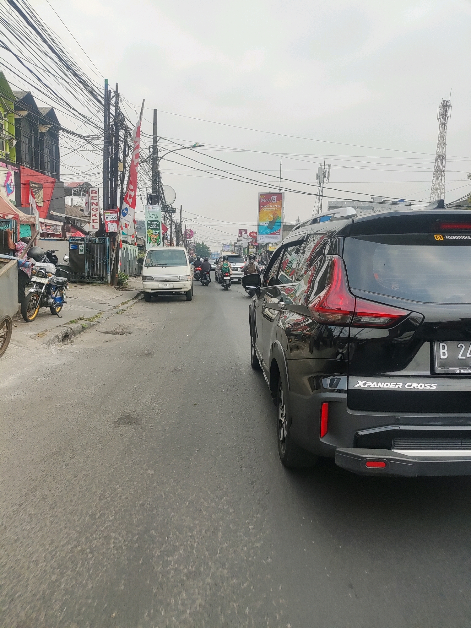 The image depicts a street scene in Bekasi, Indonesia, likely on a cloudy day. A black Mitsubishi Xpander Cross is prominently featured in the foreground, facing away from the camera. The vehicle has a license plate starting with "B 24". To the left of the Xpander, a white van is parked on the side of the road. Further down the street, several motorcycles with riders are visible, navigating traffic. The street is lined with buildings on the left side, including shops with signage in Indonesian, such as "BENGKEL" (workshop) and various advertisements. Numerous electrical wires crisscross above the street, suspended from poles. In the background, telecommunication towers can be seen. The road surface appears worn and has some patches.