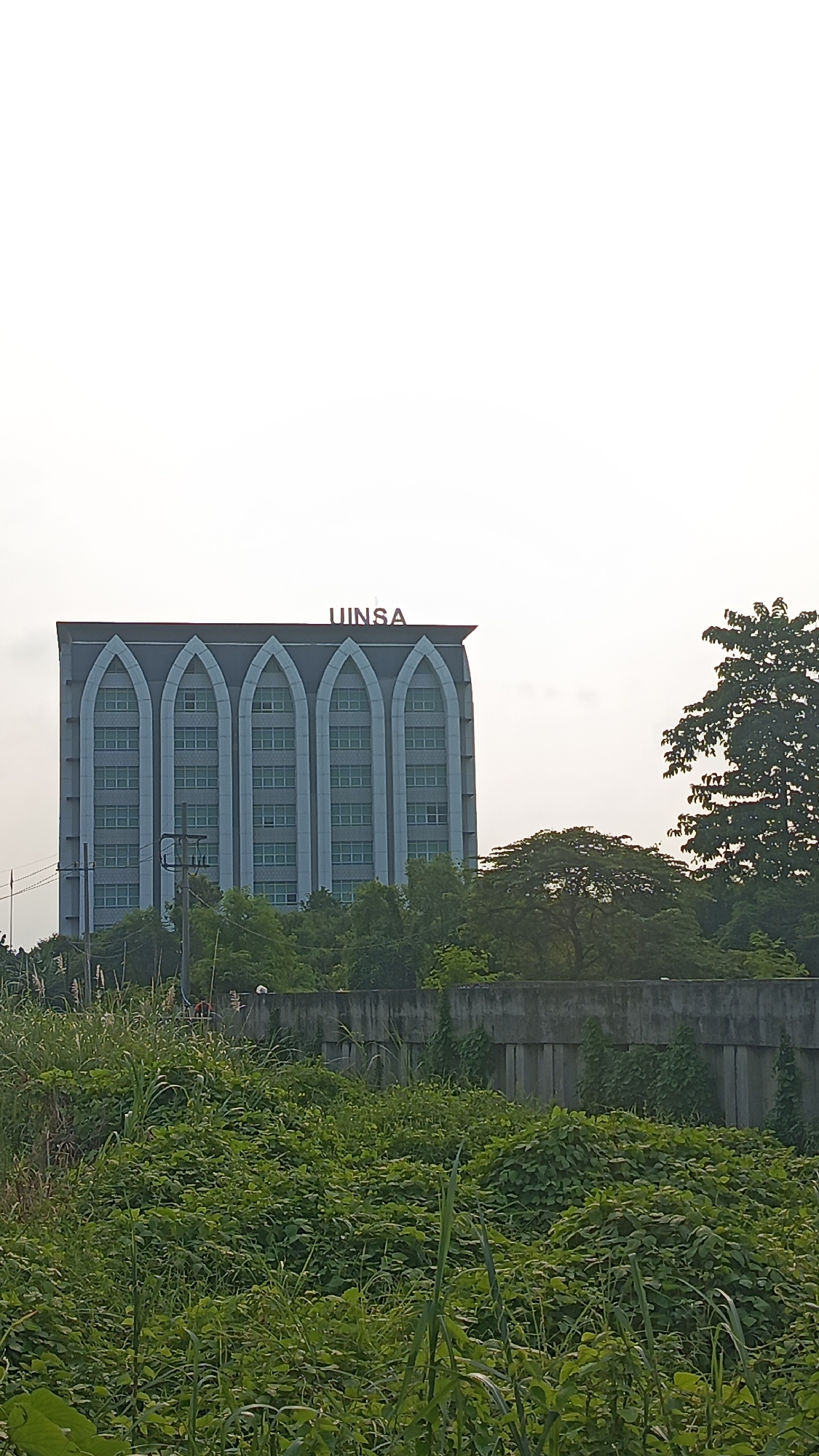 The image shows the UINSA building in Tambak Sumur, Indonesia. It's a tall building with a gray facade and arched window designs, topped with the name "UINSA" in red letters. The building is surrounded by trees. In the foreground is a field of overgrown green vegetation and a concrete wall. The sky is overcast. There are people visible in the background, behind the wall, but they are indistinct.