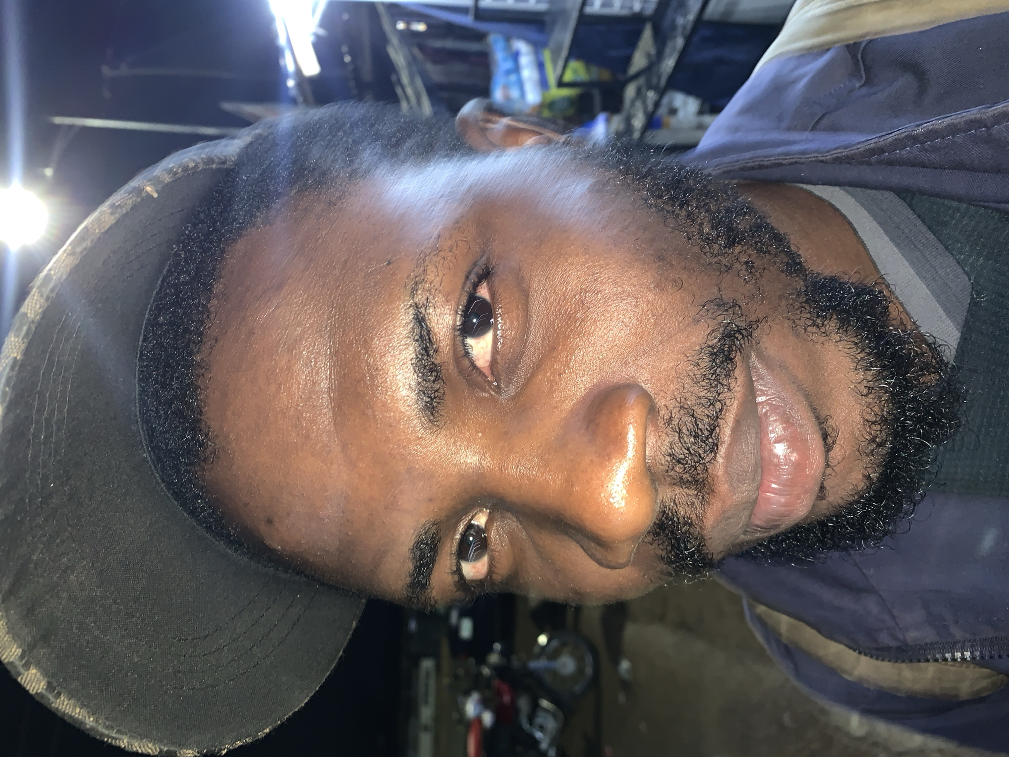 A close-up, eye-level, outdoor photograph captures a Black man from the chest up. He is wearing a dark baseball cap tilted forward, partially obscuring his forehead and hair. The man has dark skin, a full beard, and appears to be looking directly at the camera. His right eye is slightly red. He is wearing a dark blue jacket over a gray and white striped shirt. The background is blurred, but suggests an outdoor setting with some structures and possibly a motorcycle on the left. The image appears to be taken at night, indicated by the artificial lighting present in the background and the overall darkness of the scene. The photograph was taken in the city of New Stadium, Nigeria.
