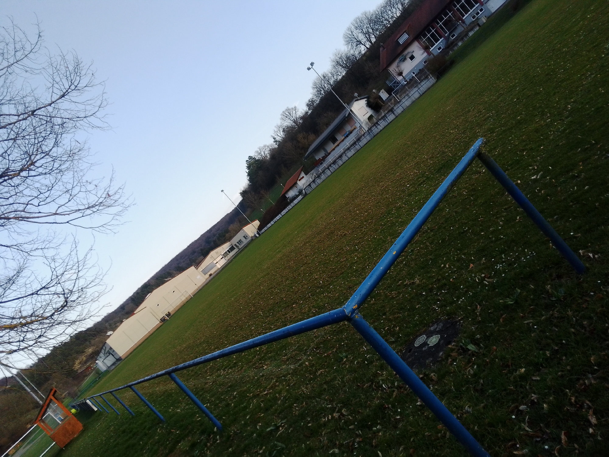 The image depicts an outdoor sports facility in Boxberg, Germany, captured from an oblique, right-tilted perspective during what appears to be late autumn or early winter.
In the immediate foreground, a weathered blue metal railing, showing signs of chipped paint and rust, occupies the lower and mid-right quadrant, forming a prominent V-shape. The ground is a patchy green grass field, sparsely covered with scattered brown leaves. Near the base of the blue railing, a dark, irregular ground patch features two light, circular markings.
Extending into the midground, another section of blue railing runs horizontally along the left edge of the field. Behind this, an orange-brown wooden utility shed with a window is positioned on the far left. Along the field's perimeter, several buildings are visible: a large, light-colored facility with a dark roof and multiple windows, and a multi-story residential-style building with a reddish-brown tiled roof situated further uphill. Tall light poles equipped with floodlight fixtures are spaced along the field's edge.
A dense treeline composed of bare deciduous trees forms the background on a hill, under a clear, light blue sky. No individuals or discernible activities are present within the frame.