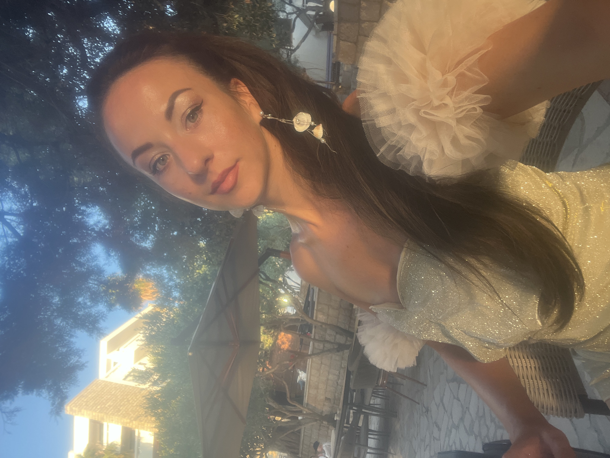 A woman is captured in a selfie outdoors, with a building and trees in the background. The woman has long, dark hair and is wearing a shimmery, light-colored strapless dress with ruffled sleeves. She is also wearing long, dangling earrings with seashell accents. The setting appears to be a patio or outdoor seating area, with tables and chairs visible. The lighting suggests it is either sunrise or sunset. The building in the background is modern in design, with white and brick facades. Trees with sparse foliage are scattered throughout the scene. The location is identified as Pečurice, Montenegro.