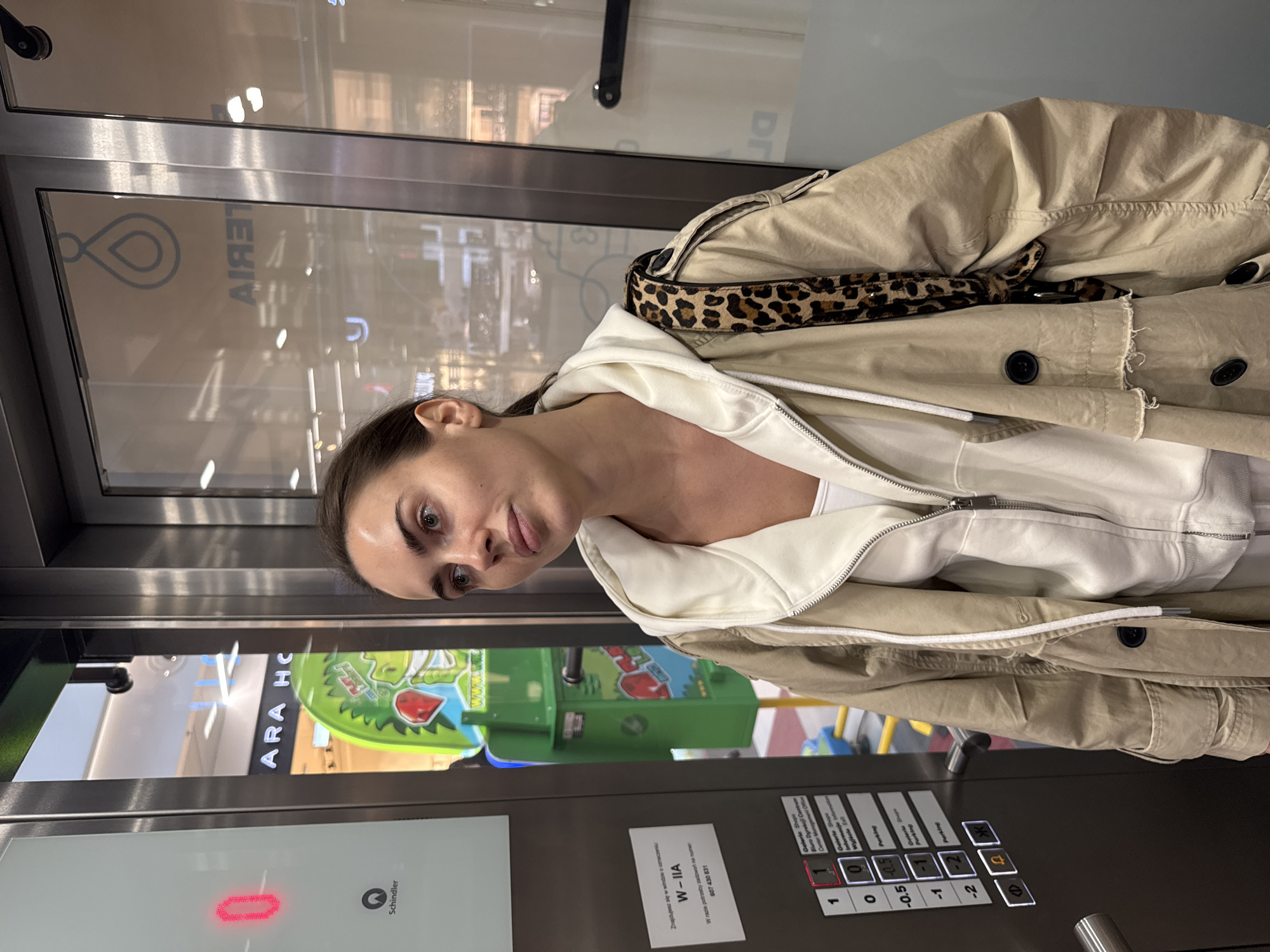 The image captures a woman inside a modern glass elevator, looking directly at the camera. She has brown hair, light-colored eyes, and a neutral expression. She is dressed in a white hooded sweatshirt layered under a beige or khaki trench coat, with a distinct leopard-print strap visible over her right shoulder.
Through the elevator's glass panels, reflections of the surrounding environment are visible, including bright overhead lights and what appears to be a colorful green children's ride or play area. Faint reflections of text like "ARA HC" and a web address fragment "www.w" are also discernible, along with a stylized loop or infinity symbol.
The elevator control panel is prominent in the lower part of the frame. It features a red digital display, potentially indicating the current floor, and numerous buttons for floor selection, including "1", "0", "-0.5", "-1", and "-2". Accompanying these buttons are labels, predominantly in Polish, such as "Galerie Shops", "Wyjście D" (Exit D), "Parking", and "Biuro Dyrekcji Centrum" (Center Management Office). The elevator manufacturer, "Schindler," is also visible.
A white sticker on the panel displays Polish text: "Znajdujesz się w windzie o oznaczeniu W-IIA. W razie potrzeby zadzwoń na numer 607 430 831," which translates to "You are in elevator designated W-IIA. In case of need, call number 607 430 831." The setting suggests a commercial building, likely a shopping mall or large retail complex, consistent with the "Galerie Shops" and "Parking" indicators. The indoor lighting is bright, and the time of day is not discernable.