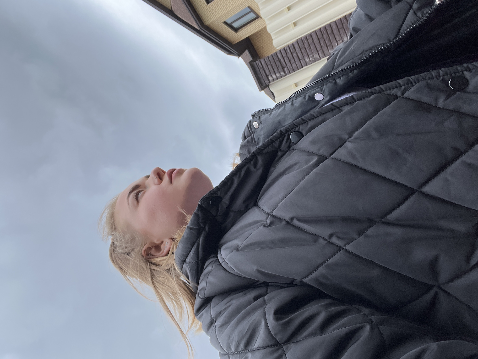 The image is a low-angle, upward shot of a young, fair-skinned person with blonde hair pulled back. They are looking upward and to the left. The person is wearing a black quilted jacket with a zipper. Above the person, there is a cloudy, overcast sky. A building is partially visible in the upper portion of the frame, constructed of beige brick with a brown roof or awning. The location cannot be forensically determined as Chaikovsky, Russia based solely on the image.