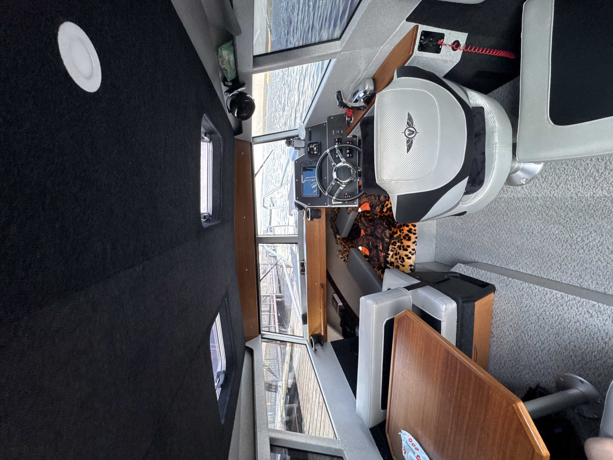 The image shows the interior of a boat docked next to a wooden pier on a body of water, likely a river or lake, under an overcast sky. The boat's cabin is equipped with a helm, a steering wheel, a throttle, a display screen showing a navigation chart, and various controls. To the left, a wooden table is set up with seating. A leopard-print blanket or cushion is visible on a seat behind the helm. The cabin has a dark upholstered ceiling with two skylights and a circular light fixture. The environment suggests a calm, potentially cool, maritime setting. There are no people clearly visible in the image.