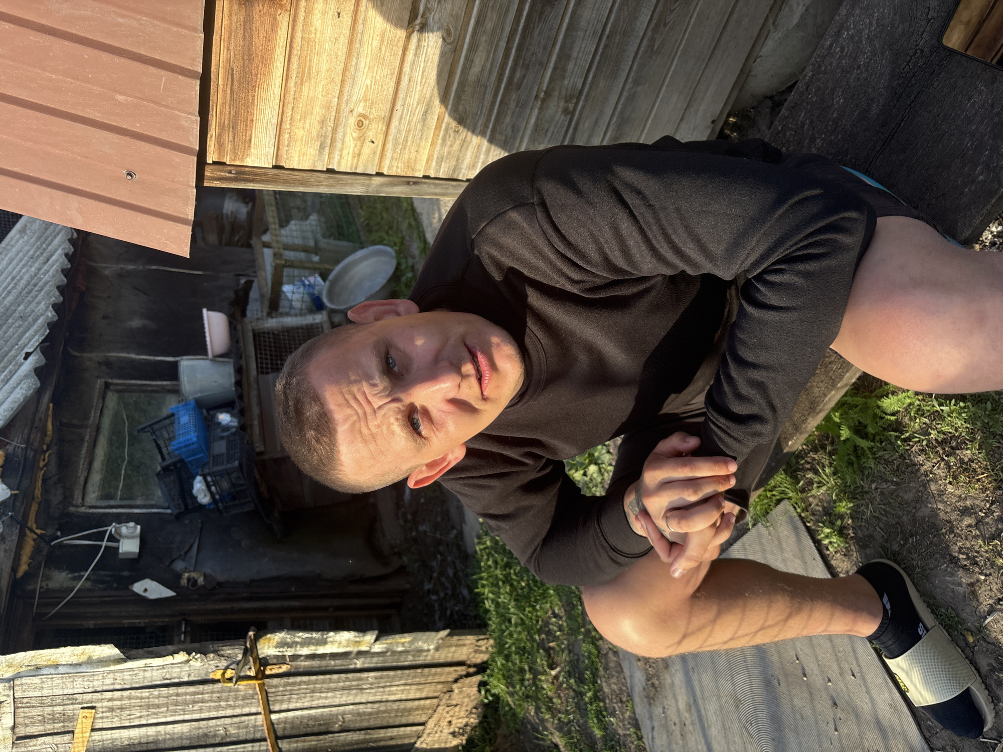A man sits on a wooden bench in front of a dilapidated wooden structure, likely in Sovkhoza "Razdol'e", Russia. The structure has a corrugated metal roof and appears to be in disrepair, with peeling paint and exposed wood. The man, with a buzz cut and blue eyes, wears a black sweatshirt and shorts. His hands are clasped, and he has tattoos on his left forearm. A ring is visible on his left ring finger. He is wearing a sandal with a black sock. The ground is covered in dirt and sparse vegetation. In the background, there are various objects, including plastic crates, a bucket, and what appears to be a metal basin. The scene is illuminated by sunlight, casting shadows.