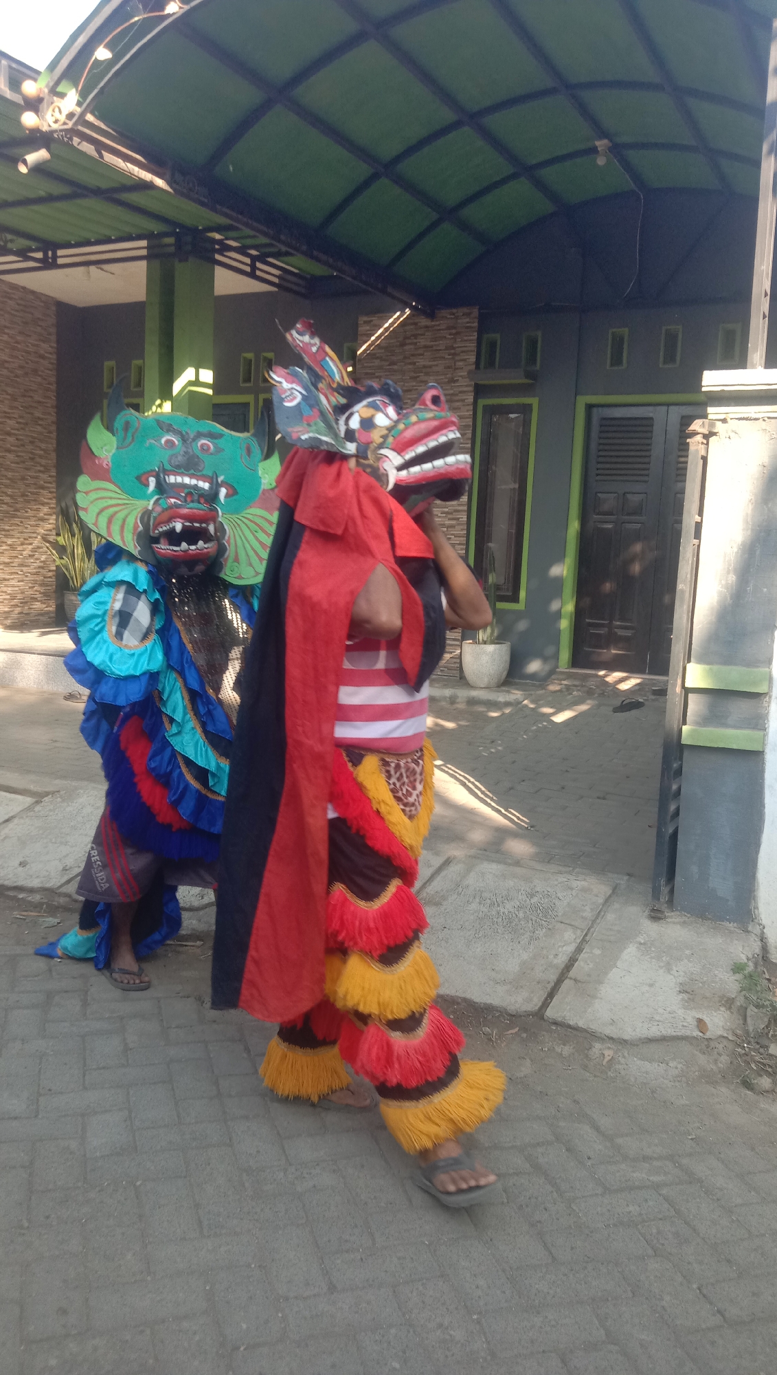 Two individuals are dressed in elaborate, colorful costumes resembling mythical creatures, possibly for a traditional performance. The person on the left wears a green and blue mask with sharp teeth and a large headdress, accompanied by a costume adorned with multiple layers of ruffled fabric in blue, red, and black. They are also wearing dark shorts and sandals. The person on the right is partially obscured by a flowing red and black cape, revealing a red and white striped shirt underneath. Their costume features a mask with a gaping red mouth and striking white teeth, and their legs are covered in layered, fringed fabric in red and yellow. They are wearing sandals. The individuals are standing on a paved walkway in front of a modern-looking building with a dark gray facade, green trim around windows and doors, and a dark green, arched roof structure overhead. The environment appears to be outdoors, in a sunny location, with a tiled pathway and some vegetation visible. The scene is likely taking place in a residential or public area in Ngempit, Indonesia, given the architectural style and cultural presentation.