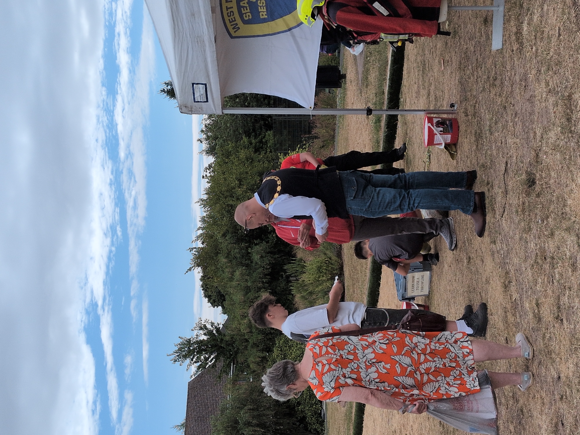 The image depicts an outdoor event on a dry, grassy field with a partially set up tent in the background. The sky is overcast with scattered white clouds. A sign on the tent reads "WEST... SEA... & RES...".
In the foreground, a woman in an orange and white floral dress stands holding a plastic bag in her left hand. To her right, a young man in a white t-shirt and dark trousers looks down at something he is holding.
Further back, a man wearing a dark vest with a gold chain and a white shirt is interacting with another person in a red jacket. A person in a grey t-shirt and dark shorts is crouching in the middle ground. To the right, a person in a red jacket and yellow helmet is partially visible, facing away from the camera.
A blue plastic container and a red bucket are on the ground near the tent. The overall setting suggests a community event or demonstration. The city of Dawley, United Kingdom is mentioned as the location.