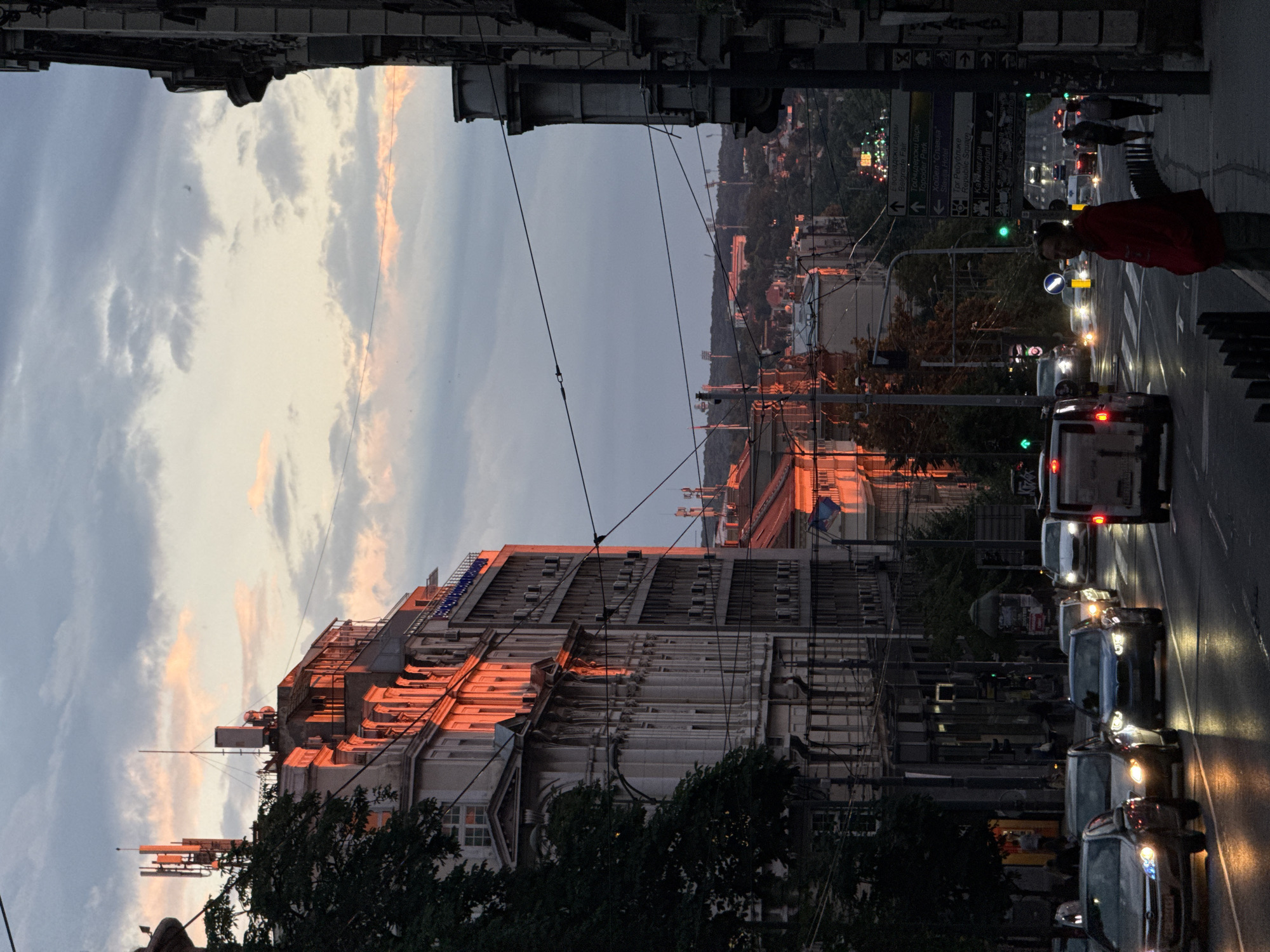 The image depicts a street scene in the Vračar urban municipality of Serbia during sunset. The warm, golden light of the setting sun illuminates the facades of buildings on the left side of the street, highlighting architectural details. The sky is filled with a mix of gray and pinkish clouds. A street with several lanes of traffic is visible, with cars driving and their headlights and taillights reflecting on the wet asphalt, suggesting recent rain. On the right side of the street, the darker facades of buildings are visible, along with streetlights and traffic signals. Pedestrians are present on the sidewalk, and one man in a red jacket stands out. Electrical wires crisscross overhead, typical of an urban environment. In the distance, a cityscape with more buildings and hills can be seen, with stadium lights visible on a distant hill. Signs in Cyrillic script are posted at an intersection, indicating street names or directions. The overall impression is a bustling urban environment at the end of the day, bathed in the dramatic light of sunset.