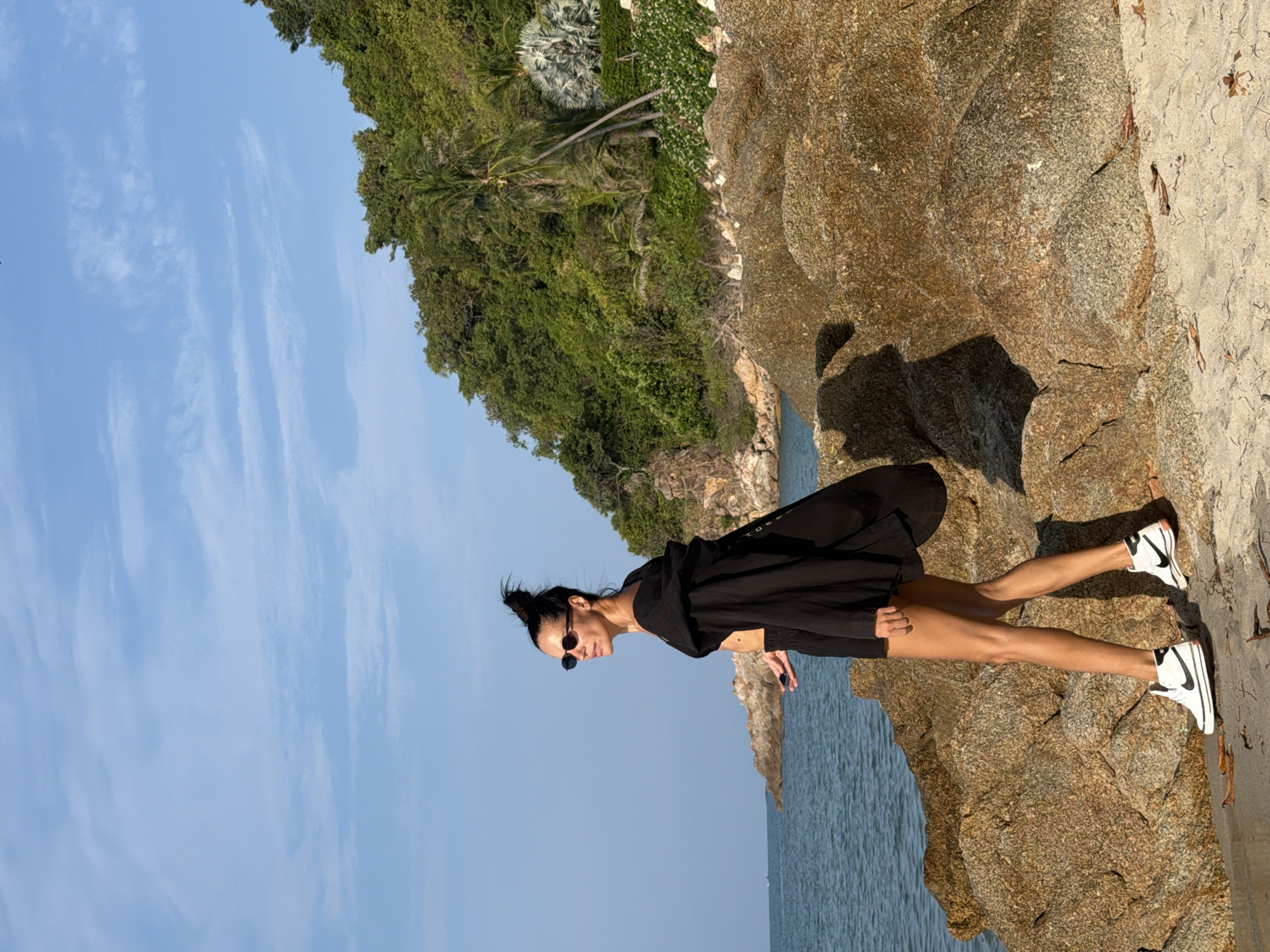 In Ko Samui District, Thailand, a woman stands on a rocky beach facing the ocean. She has dark hair pulled up in a bun and wears sunglasses, a black top, black shorts, and a flowing black open jacket. Her left hand is outstretched, holding a small, dark object. She is wearing white sneakers with black accents. The background consists of the turquoise ocean, a lush, green, tree-covered hillside, and a clear blue sky with wispy clouds. The rocks in the foreground are large and brownish-gold.