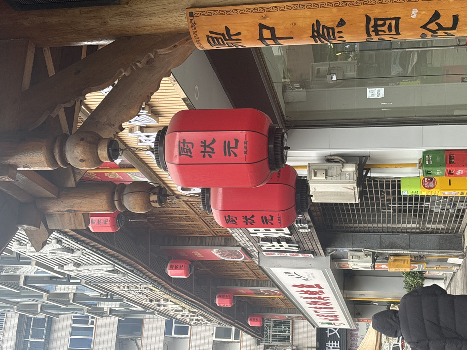 This image depicts a street scene in Jiangjin, China, under an overcast sky. In the lower-left, a single individual, wearing a black hooded puffer jacket, walks away from the camera on a grey concrete sidewalk. The foreground is dominated by a building facade featuring a substantial wooden overhang with carved supports and decorative elements. Suspended from this overhang are multiple red cylindrical lanterns. Two large lanterns in the immediate foreground display vertical black Chinese characters that translate to "Chef Champion." To the far right, a vertical wooden plank affixed to a structural pillar bears black Chinese characters translating to "Discussing wine brewed in a tripod."
Below the wooden overhang, various commercial storefronts are visible. One storefront to the left features a white rectangular sign with red Chinese characters. Adjacent to it, another storefront is secured by a pulled-down corrugated metal roller shutter, with a sign above reading "Fat Sister" in Chinese characters. Several wall-mounted outdoor air conditioning units are affixed to the building facades, along with vertical yellow utility pipes. On the sidewalk in front of the closed storefront, a stack of colorful plastic crates (red, green, yellow) and a yellow rectangular utility box are present. In the upper-left background, a multi-story residential or commercial building with numerous windows is visible. A portion of a white tent structure is partially observable on the far-left edge of the frame.