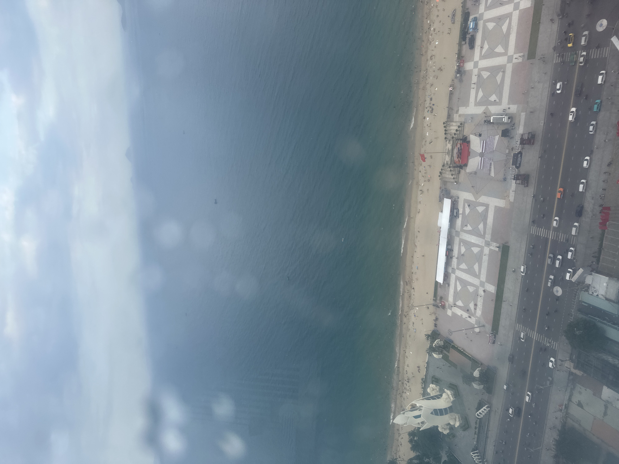 This high-angle, aerial photograph presents a panoramic view of a bustling coastal area, consistent with Nha Trang, Vietnam, under bright, possibly hazy, daytime conditions. The scene is organized into distinct horizontal bands: the sea, a sandy beach, a wide promenade, and an urban road lined with buildings.
The leftmost portion of the image features a calm, blue-green ocean with subtle surface ripples. Adjacent to the ocean is a sandy beach, densely populated with numerous people, umbrellas, and what appear to be beach chairs. Several red flags are visible along the shoreline.
Next is a broad promenade or plaza, distinguished by its light-colored paving and prominent grey and white geometric patterns. This area is also active with pedestrians. Notably, several temporary structures are present, including large white tents and a stage-like setup with a red canopy, suggesting a public event or festival is either underway or recently concluded.
To the right of the promenade lies a multi-lane asphalt road, busy with vehicular traffic, primarily cars and motorcycles. White road markings, including lane dividers and crosswalks, are visible. On the far right, beyond the road, various urban buildings are visible, including a distinctive white, futuristic-looking structure with curved architecture in the lower right corner. The sky on the far left shows some cloud cover or haze. No explicit text is clearly discernible from this high vantage point.