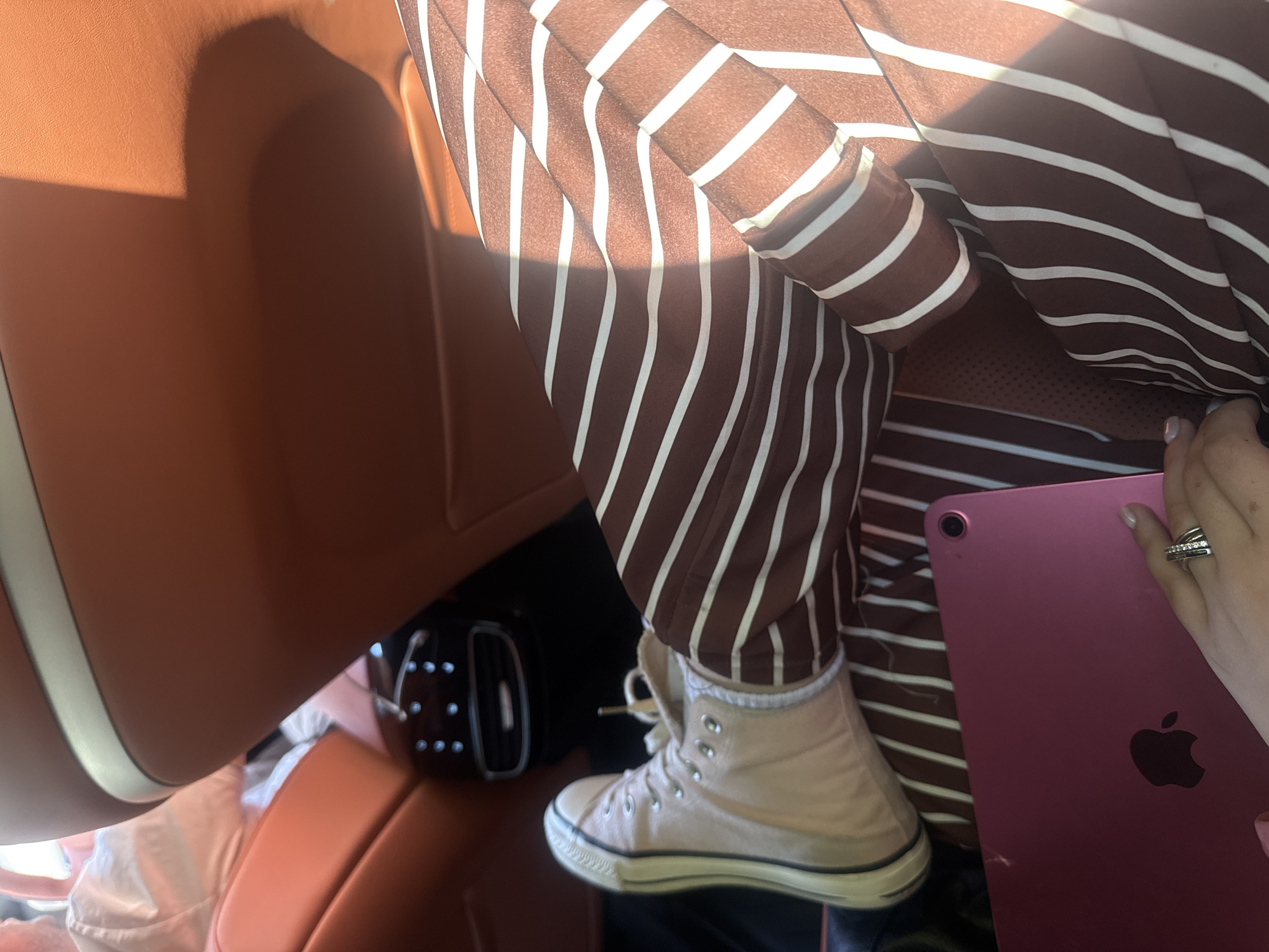 In the interior of a vehicle, a person's legs are visible, dressed in brown and white striped pants and light-colored high-top sneakers. A pink tablet rests on their lap. The background shows the brown leather interior of the car, including a headrest and a portion of a vent. The scene is illuminated by sunlight, casting shadows. No specific location in Yerevan, Armenia is identifiable from the image.