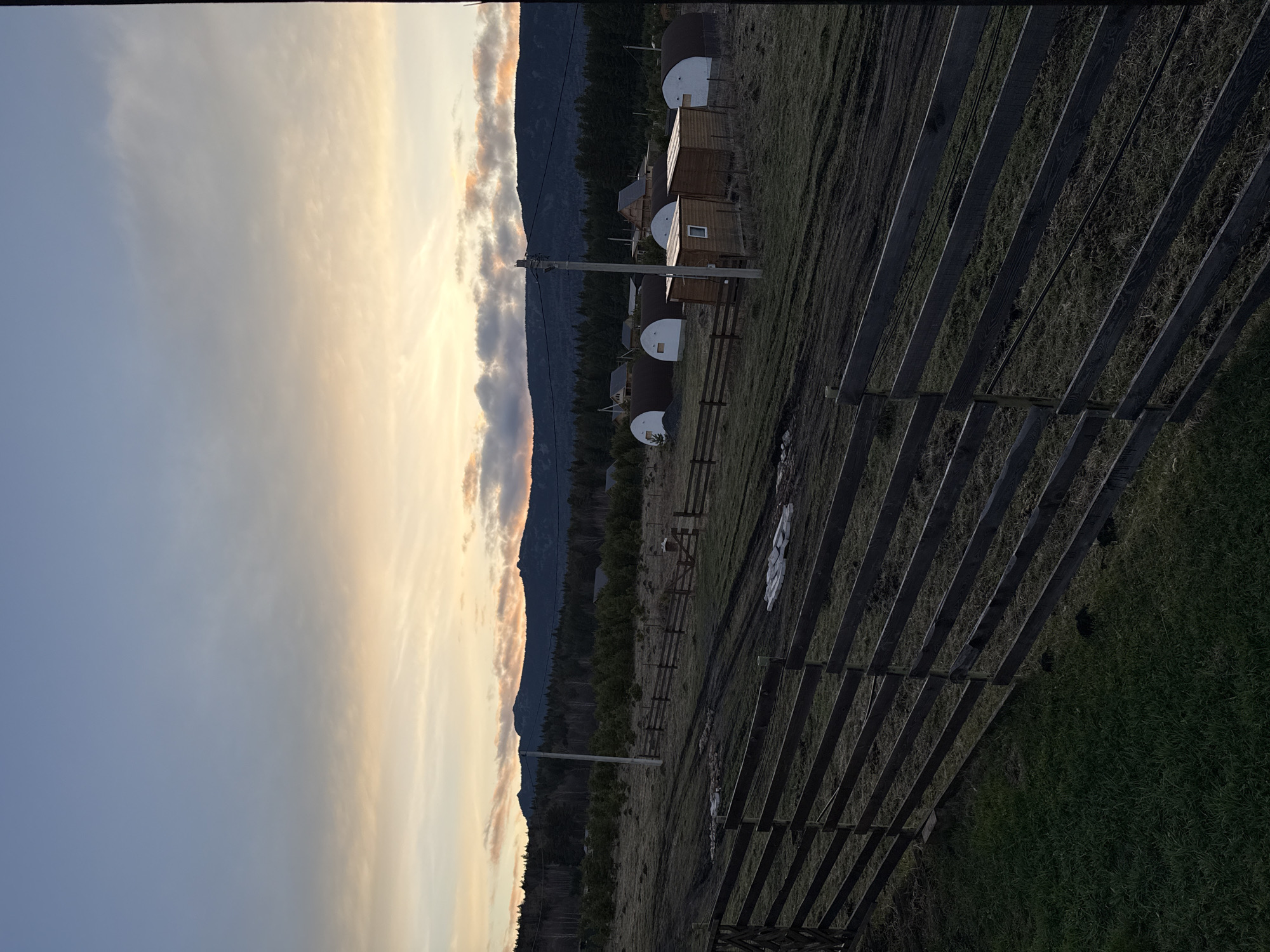 This image captures a serene rural landscape in Tyulyuk, Russia, likely during sunrise or sunset. The sky displays a striking gradient, transitioning from a soft blue on the left to warm, vibrant hues of orange, yellow, and pink towards the right, indicative of twilight. Scattered clouds are visible, some catching the warm light while others appear darker.
In the background, a dark, forested mountain range or series of hills forms the horizon line. The mid-ground features a cluster of small structures, including several distinct, rounded, dome-like buildings with white tops, interspersed with rectangular wooden cabins, some equipped with small windows. Utility poles with overhead wires are also present, connecting these dwellings.
The foreground is dominated by a grassy field, which appears somewhat dry with patches of greener grass, particularly towards the lower right. A dark wooden fence with multiple horizontal rails runs diagonally across the right side of the image, drawing the eye into the scene. On the ground in the mid-distance, some white tarps or coverings are visible. A faint, indistinct animal, possibly a horse or cow, can be seen in the distance near the fence line.
The overall scene is tranquil and devoid of overt human activity, suggesting a peaceful, rustic setting, possibly a glamping site or a remote village accommodation nestled within a natural, mountainous environment. There is no visible text in the image.