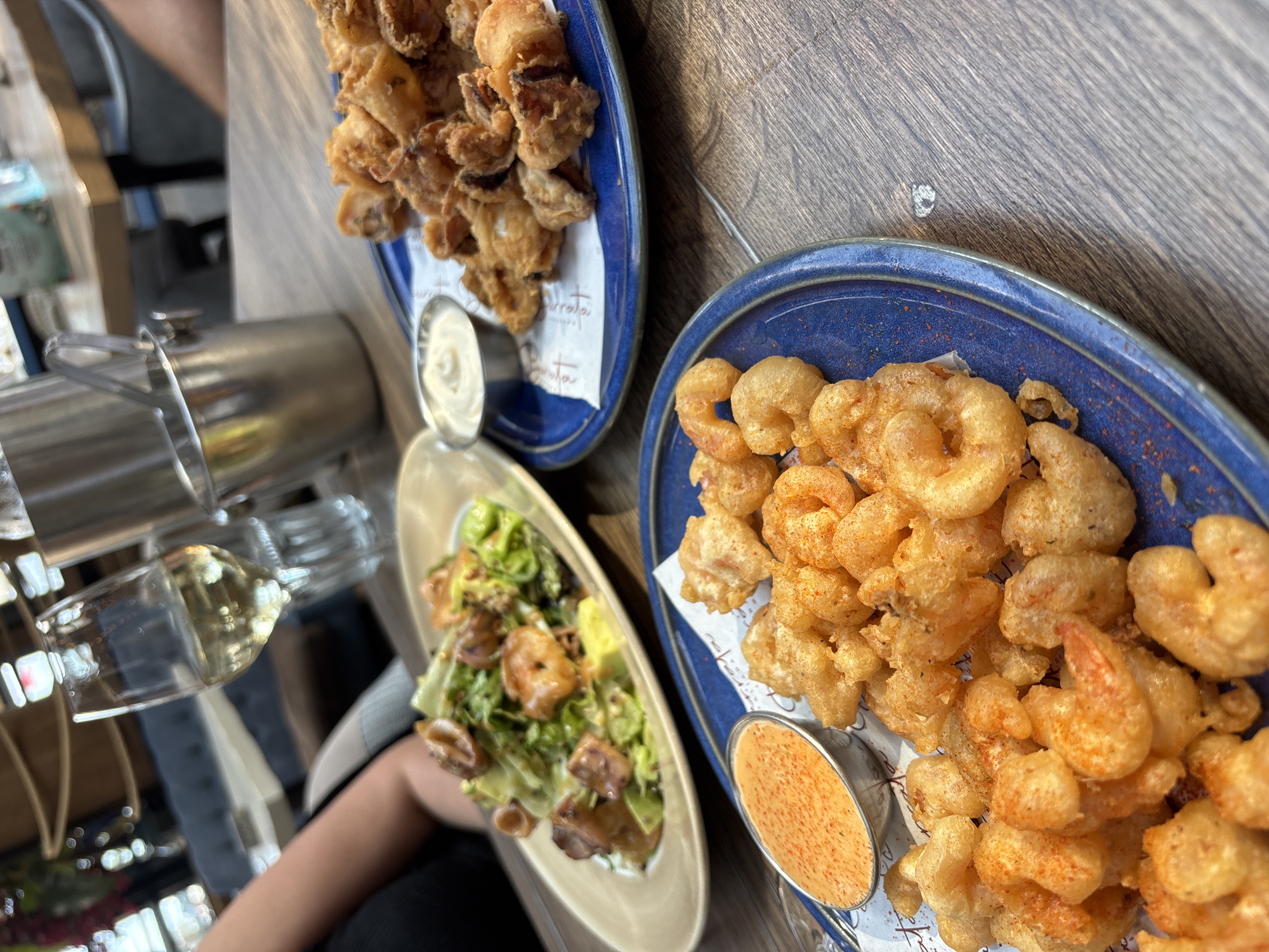 The image presents a top-down view of a dining table set with multiple seafood dishes and beverages, indicative of a restaurant meal. The table surface is a dark, wood-grained material.
Prominently featured are two large plates of fried seafood. In the upper central area, a blue ceramic plate contains a generous portion of golden-brown fried calamari rings, appearing crispy. These rest on a white paper liner that displays partially visible, stylized text, which seems to read "Duarata" or a similar word, possibly a restaurant name or brand. Accompanying this dish is a small silver cup holding a creamy white dipping sauce.
To the right, another blue ceramic plate is filled with an abundance of golden-fried shrimp. These shrimp also appear crispy and are lightly dusted with a red seasoning. A small silver cup containing an orange-red dipping sauce, also sprinkled with red seasoning, sits beside the shrimp.
Further down and to the left, an oval-shaped beige plate holds a fresh salad. This salad features green leafy vegetables, what appears to be grilled or sautéed shrimp, and possibly other ingredients like croutons or avocado.
On the far left, a clear wine glass filled with a light-colored liquid, presumably white wine, is visible. Beside it, a metallic pitcher or carafe stands. A person's arm, dressed in a black sleeve, is partially seen at the bottom left, suggesting someone is seated at the table. The background is softly blurred, consistent with an indoor restaurant environment. The scene captures a convivial and casual dining experience centered around seafood.