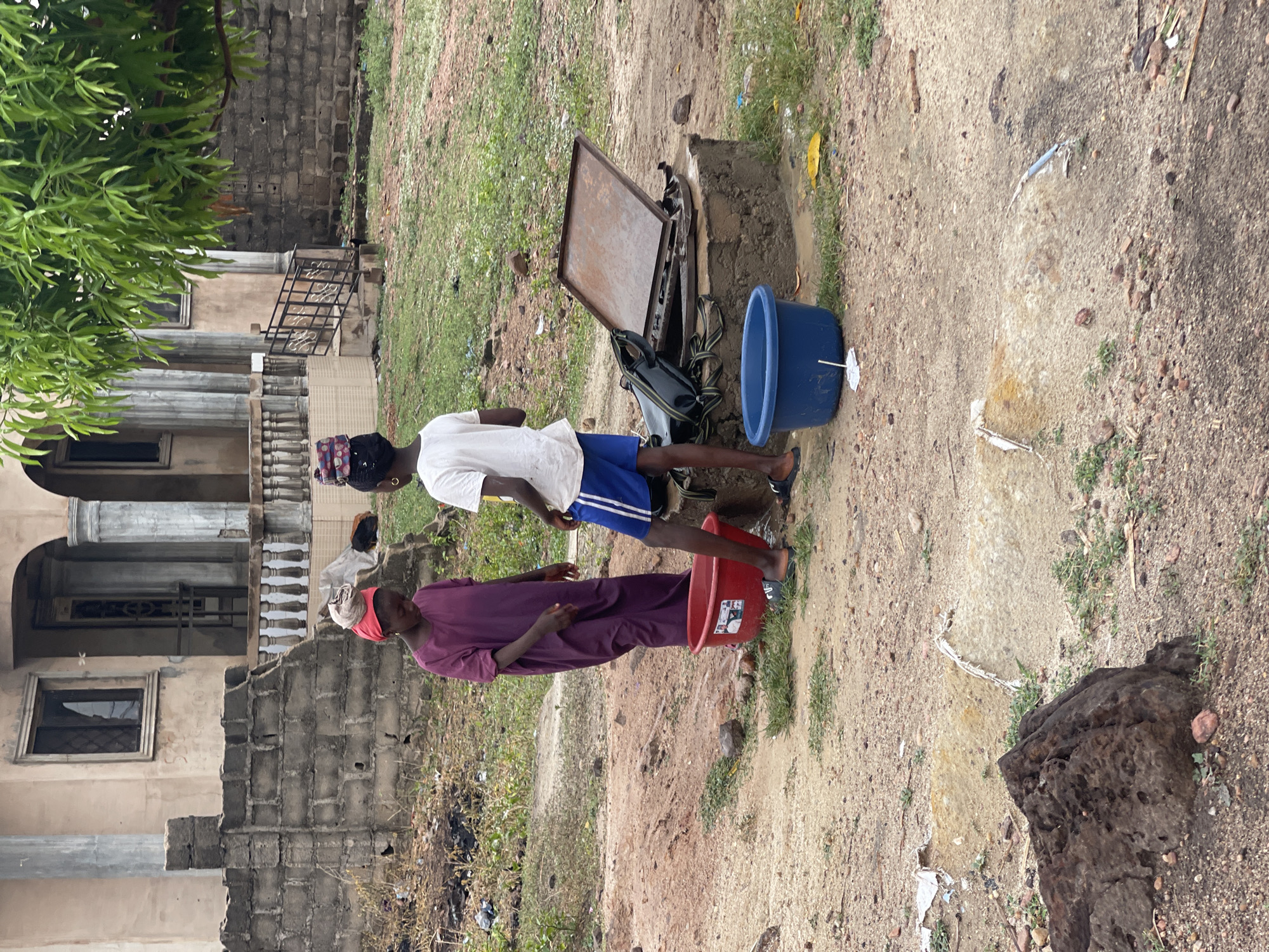 Two people stand outdoors near a concrete structure with an open metal lid. The person on the left is wearing a maroon robe and red head covering, standing beside a red bucket. The person on the right is wearing a white shirt and blue shorts. A blue bucket sits next to the concrete structure. The area is a dirt and gravel surface, with grass patches and buildings in the background, likely somewhere in the city of Lokoja, Nigeria. The actions appear to be related to the water in the buckets and the water from within the concrete structure.