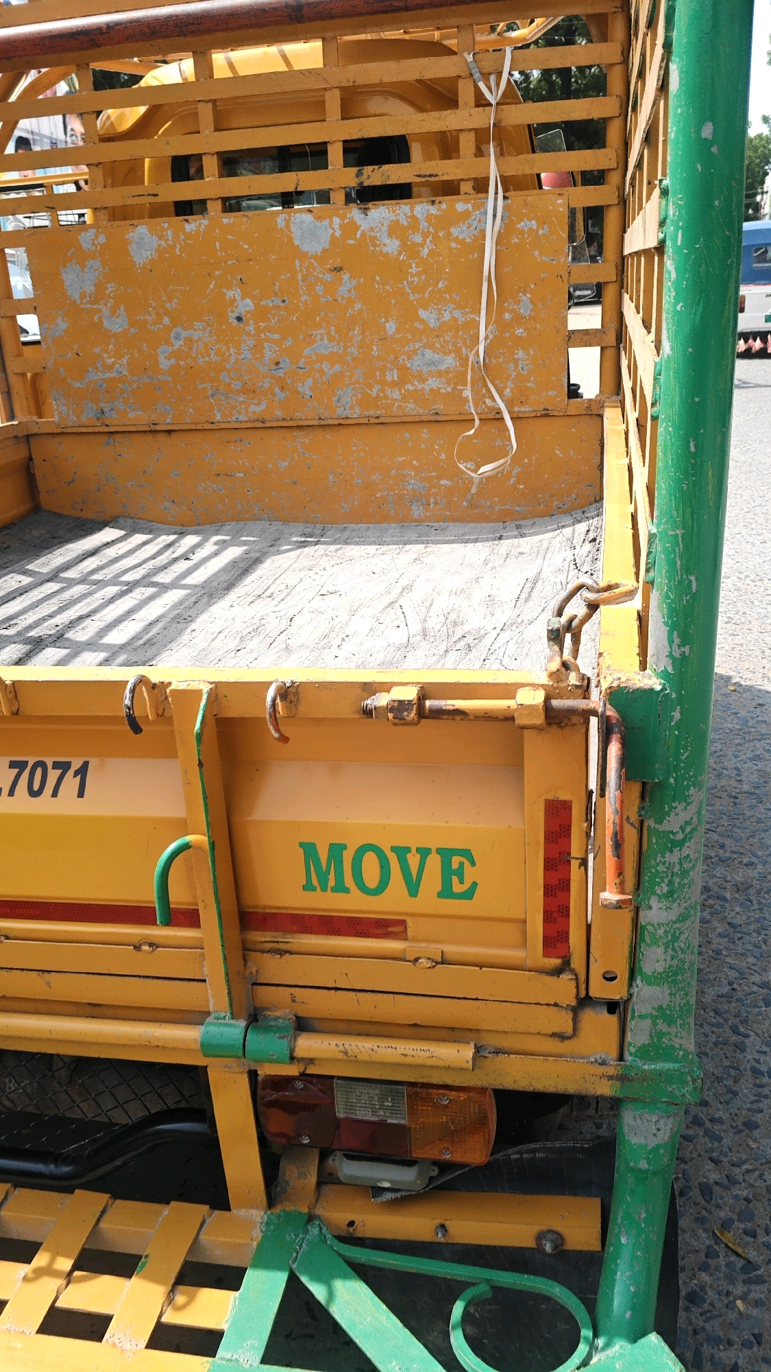 The image shows the rear of a yellow, open-bed three-wheeled vehicle, possibly a tempo or auto-rickshaw, parked on a paved road. The vehicle's cargo area is partially visible, with a gray, dusty floor. A white ribbon is tied to the upper railing of the vehicle and hangs down. The side of the vehicle has the word "MOVE" painted in green. There is also some lettering with the number "7071" visible. Red and white reflective tape is present on the lower rear section of the vehicle. A green metal bar runs vertically along the right side of the vehicle, and a tail light assembly is mounted below. The background shows a blurred view of trees and buildings, suggesting an urban or semi-urban setting. The overall impression is of a functional, somewhat weathered utility vehicle.
