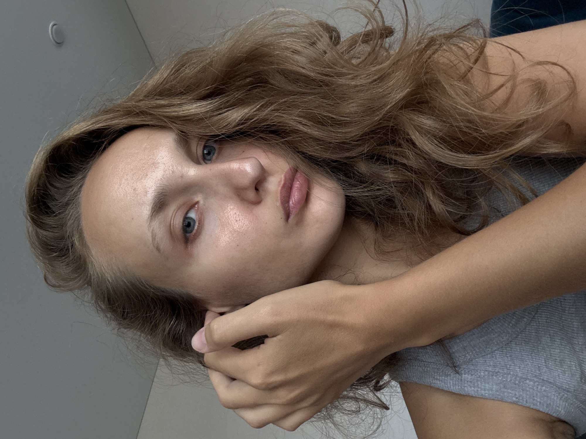 A medium shot captures a young woman indoors, her face partially obscured by her hand as she adjusts her hair near her ear. She has light brown, wavy hair and blue eyes. Her skin appears fair with some visible texture. She is wearing a grey tank top. The background consists of a plain wall and a ceiling. The image is well-lit, likely with natural light.