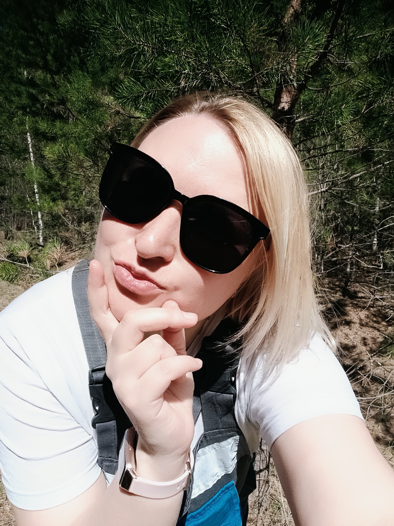 A woman poses for a selfie outside. She has blonde hair, wears large black sunglasses, a white t-shirt, and a jumpsuit with blue accents. Her hand is raised near her chin in a thinking pose. She wears a light pink smartwatch. Behind her is a forest with pine trees. The location is likely in or around the city of Chaikovsky, Russia.