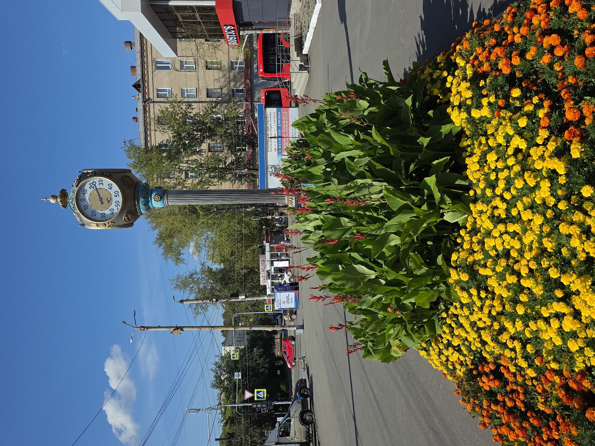 The image shows a street scene in Chelyabinsk, Russia, on a sunny day. In the foreground, there is a large, brightly colored flowerbed featuring yellow and orange marigolds, with tall green plants and red flowers behind them. A classic ornate clock with a thermometer is prominently displayed in the mid-ground, mounted on a decorative pillar. Behind the clock, a multi-story brick building with many windows is visible, along with trees and a modern building with a red and grey facade and the sign "ROSTIC'S". Red buses are parked in front of the modern building. Several cars, including a white van and a red car, are on the paved road, and pedestrian crossings with yellow warning signs are also visible. Power lines stretch across the bright blue sky with a few scattered white clouds. The overall impression is a well-maintained urban environment with floral decorations.