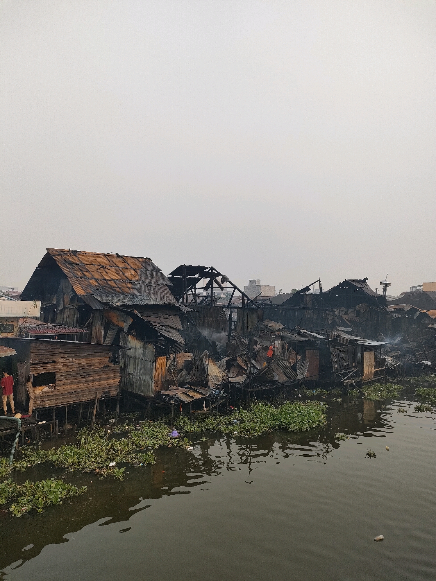 The image depicts a section of a stilt village in Banjarmasin, Indonesia, that has been devastated by a fire. The structures, made of wood and corrugated metal, are heavily charred and appear on the verge of collapse. Smoke is still visible rising from some of the damaged buildings, indicating that the fire may have recently occurred or is still smoldering. The buildings are situated along a narrow waterway, which is covered in aquatic vegetation and debris. In the foreground, a person wearing a red shirt is standing on a wooden platform. The sky is overcast and the overall atmosphere is somber, suggesting the aftermath of a significant disaster.