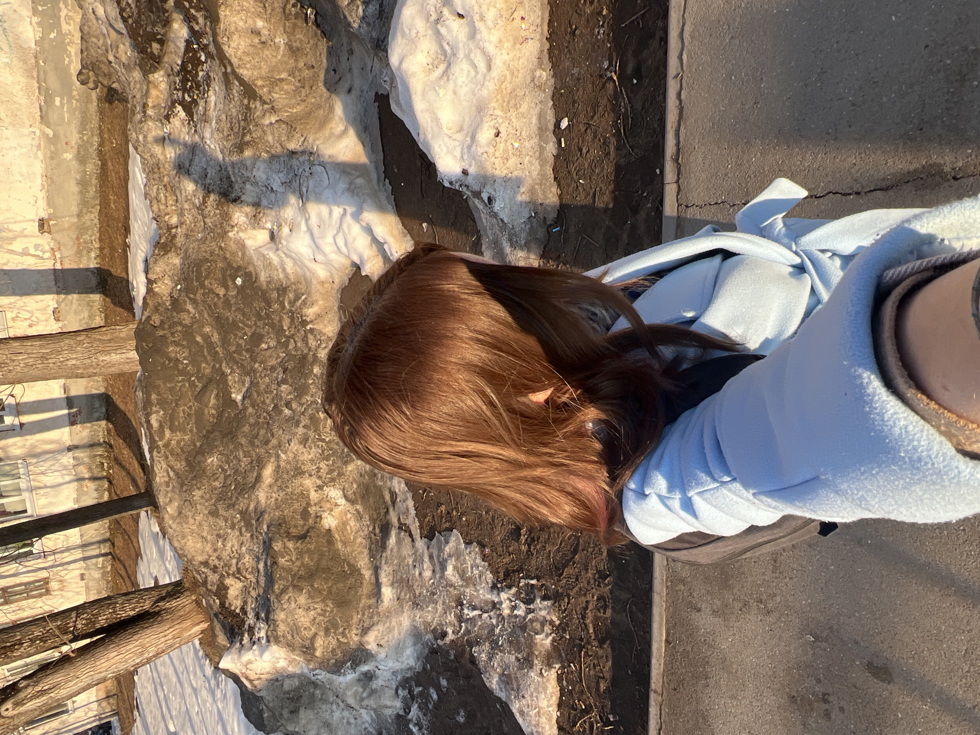 A high-angle selfie captures the upper torso and head of a person with long, reddish-brown hair, partially obscuring their face. The individual wears a light blue, soft-textured garment, possibly a hooded jacket or scarf, over a darker layer, with a beige cuff visible at the bottom. The scene is set in an urban or suburban environment within Chaikovsky, Russia, characterized by early spring conditions. The ground features a grey asphalt surface, transitioning into an area of patchy, dirty, melting snow and dark, exposed earth containing scattered debris, including small white and blue fragments and cigarette butts. Several bare-trunked trees stand in the midground, casting long, distinct shadows across the snow and ground. In the background, a weathered, light-colored building with multiple windows displays discoloration and signs of aging on its facade. The scene is illuminated by low-angle sunlight, indicative of morning or late afternoon.