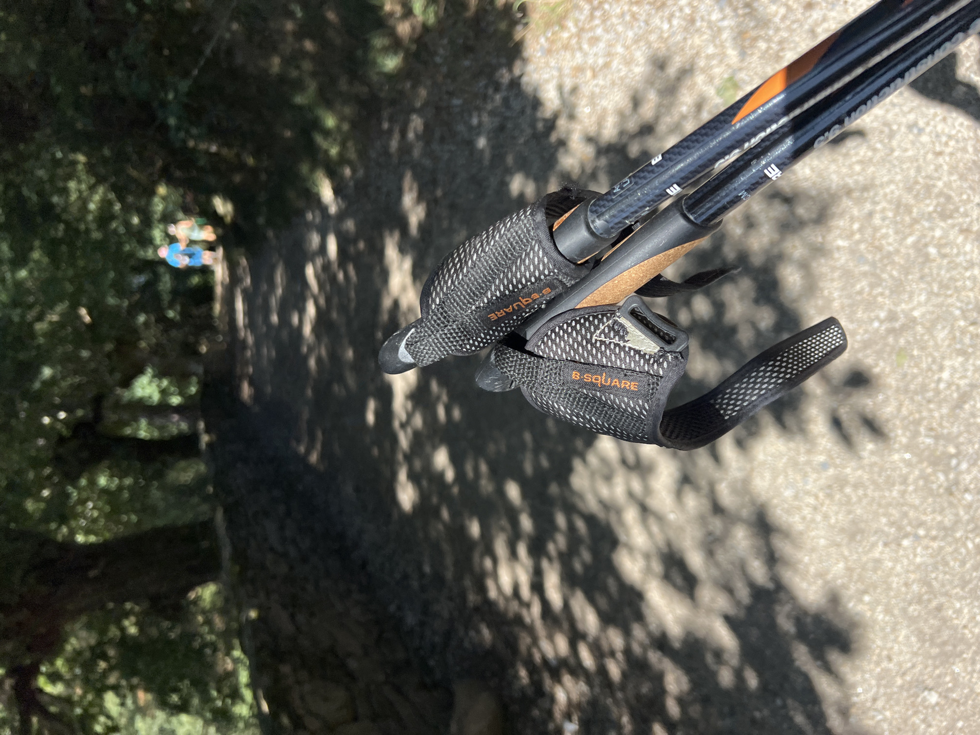 The image shows a pair of trekking poles with black and orange accents in the foreground, held by an unseen person. The poles are positioned diagonally across the frame, with the grips and straps clearly visible. The background depicts a sun-dappled dirt path, likely in a wooded area. Two blurry figures, appearing to be walking away from the camera, are visible in the distance on the path. The environment suggests a rural or natural setting, possibly a trail for hiking or walking. The lighting indicates it is daytime, with strong sunlight casting shadows from the trees onto the path. The location is identified as Paradela, Spain.