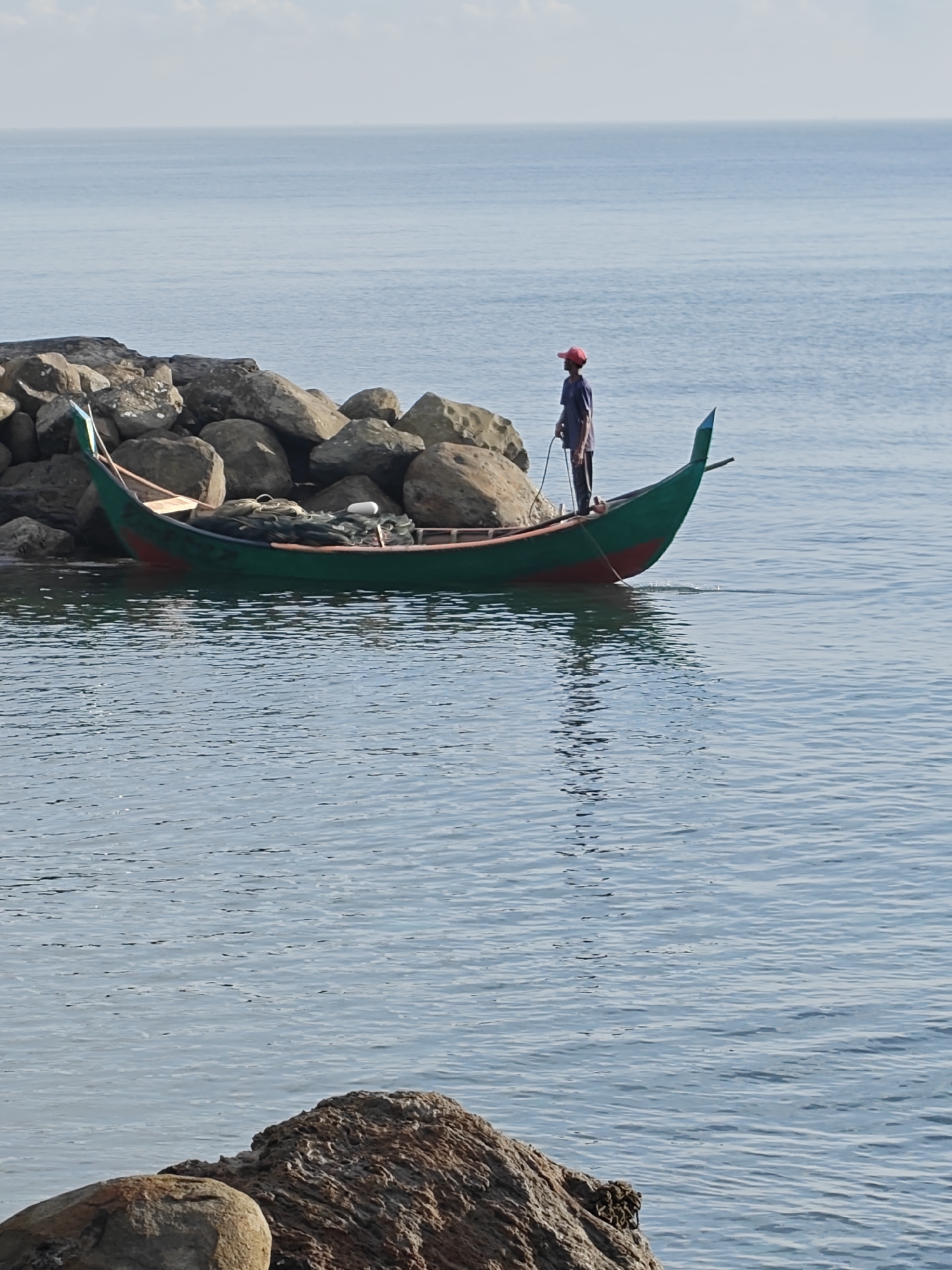 In a serene marine scene, a slender green and red wooden boat rests partially beached against a rocky breakwater along the coast of Lhokseumawe, Indonesia. Inside the boat, fishing nets and other gear are visible. A man wearing a red cap and a dark shirt stands in the boat, holding what appears to be a rope or tether connected to the shore. The water is calm, reflecting the boat and the man. The backdrop is the tranquil ocean meeting the horizon under a lightly clouded sky. Rocky formations are also visible in the foreground.