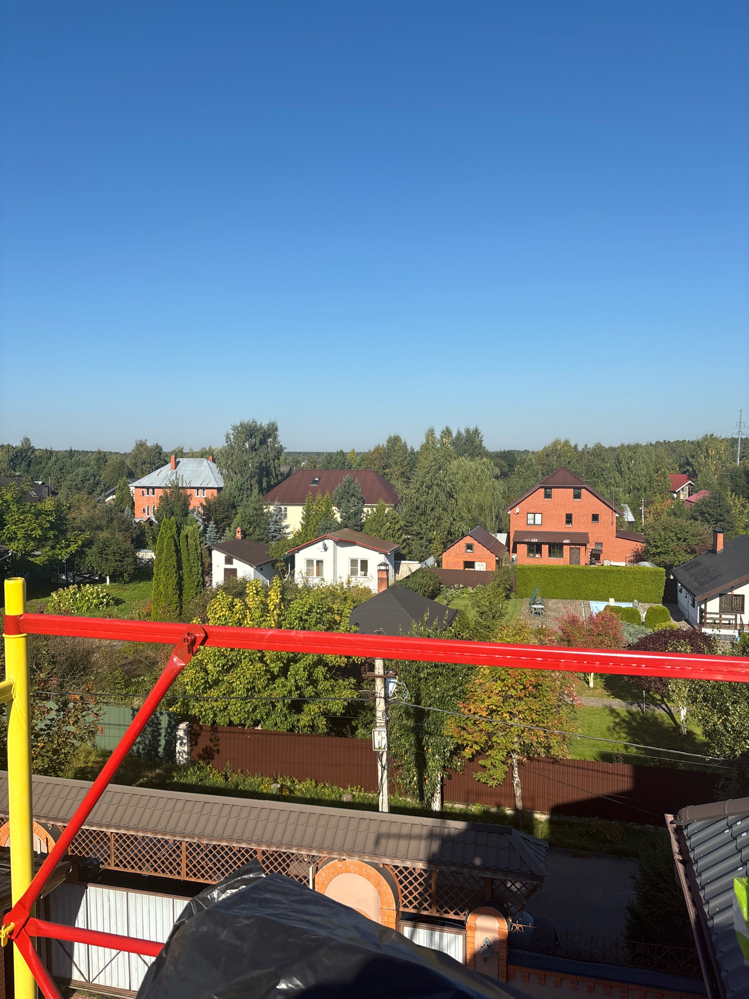 The image captures a suburban residential area on a clear, sunny day. The foreground features scaffolding with yellow and red poles, suggesting ongoing construction or maintenance. Beyond the scaffolding, a cluster of houses is visible, varying in architectural style and size. Some houses are brick, while others are painted white. They are nestled among lush green trees and manicured lawns. The sky is a vibrant, cloudless blue. The overall impression is one of a peaceful, well-kept neighborhood. The image is believed to have been taken in the city of Zhavoronki, Russia, given the architectural styles and vegetation consistent with the region.