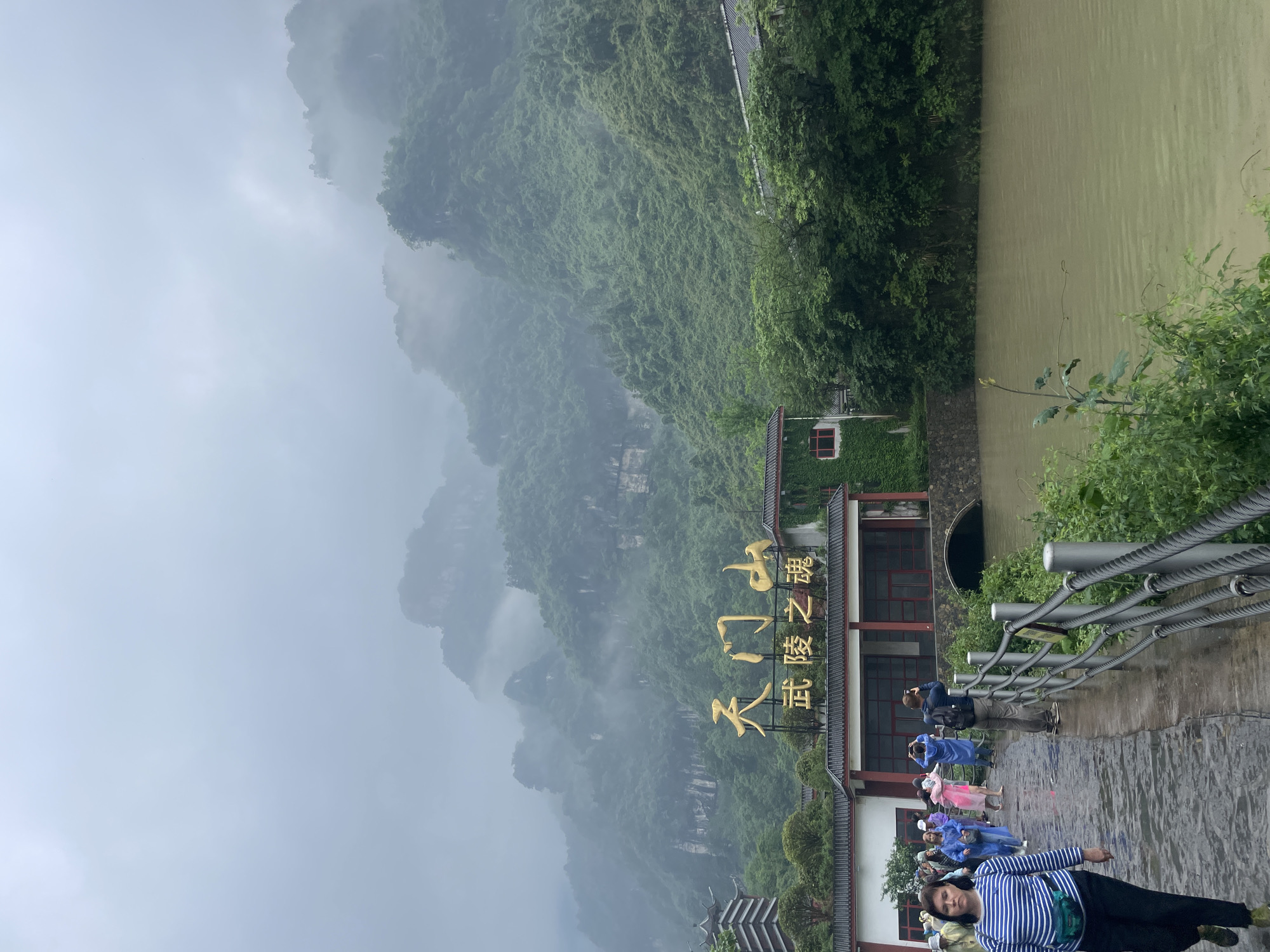 The image depicts Tianmen Mountain in Yongding District, China. The mountain is lush with vegetation and shrouded in mist. In the foreground is a building with a sign in gold Chinese characters. People are standing on a pathway in front of the building, some of them taking photos. A body of water is visible to the right. The weather appears overcast and rainy.