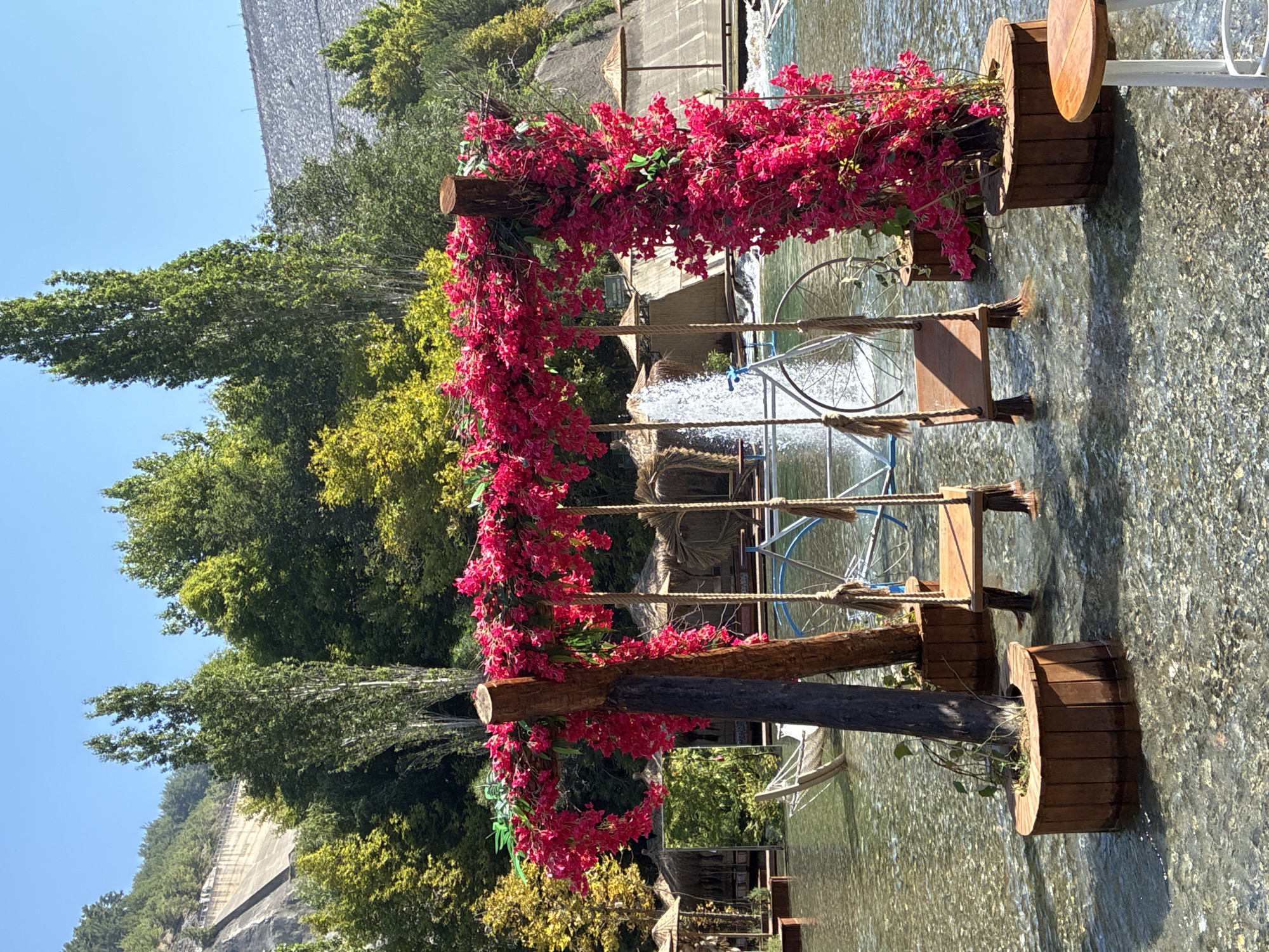 The image captures a scenic outdoor setting, likely a cafe or resort, with a focus on a decorative swing set immersed in shallow, clear water. The swing set is constructed from natural wood and adorned with vibrant pink bougainvillea flowers cascading from an overhead archway. Two wooden seats hang from ropes, partially submerged in the water. In the background, a waterfall feature adds to the ambiance, and a vintage bicycle with large spoked wheels is positioned behind the swings. Several thatched-roof cabanas and umbrellas are visible, suggesting a relaxation or dining area. The surrounding environment is lush with trees and vegetation, and a large concrete dam structure is prominent on a hillside in the distance. The water appears to be a natural or artificial stream flowing through the area, with a rocky, clear bottom. The sky is a bright, clear blue, indicating fair weather. The location is Alanya, Turkey.