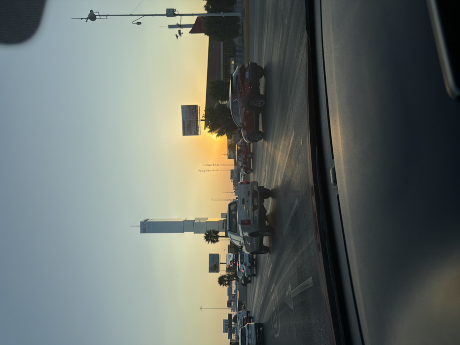 The image captures a road scene through a car window. Multiple vehicles are in traffic on a road lined with palm trees and advertising signs. A tall building is visible. The sky is bright, suggesting either sunrise or sunset. The view is taken from inside a vehicle, focusing on the outside traffic conditions.