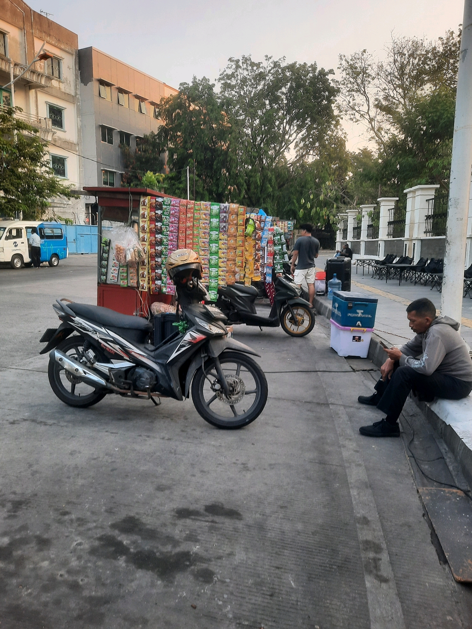 The image captures a street scene in North Jakarta, Indonesia, with a focus on a stall selling snacks and drinks. In the foreground, a black motorcycle is parked, with a yellow helmet on its handlebars. To the right, a man sits on a concrete ledge, looking at his phone. Behind him, a blue cooler box and a water jug are visible. Further back, a stall displays a wide variety of packaged snacks and drinks. Two individuals are seen near the stall, one appearing to be a vendor and the other a customer. In the background, a building with multiple floors and trees suggest an urban environment. The lighting indicates it is likely late afternoon or early evening.