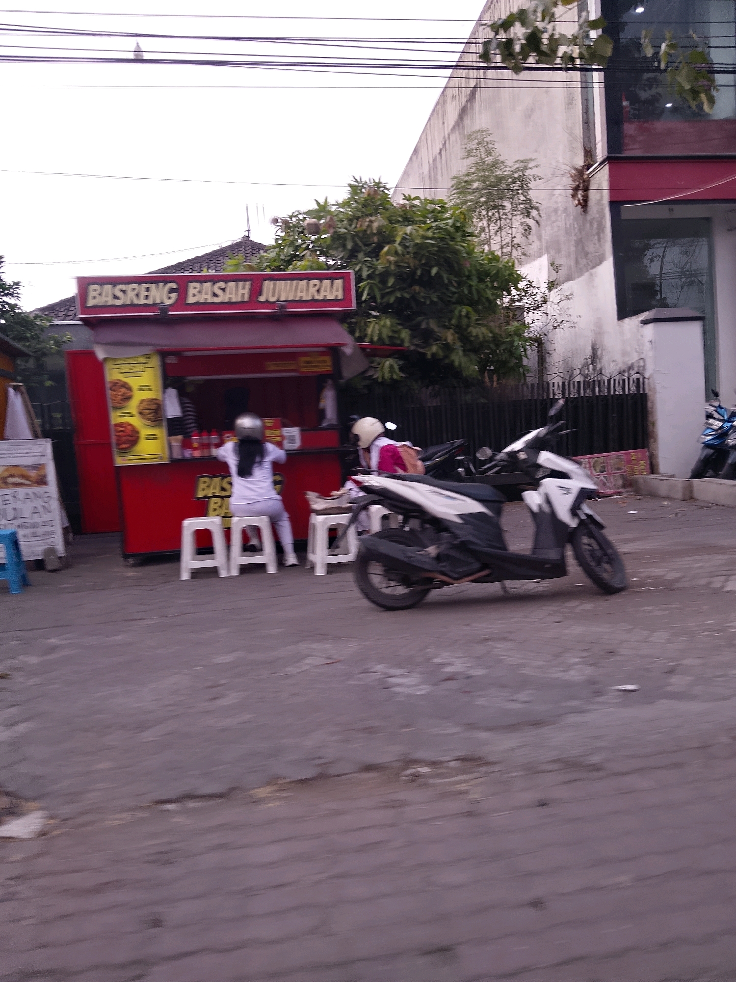 A street-level, medium shot captures a red food stall in Kesatrian, Indonesia, with a white and black scooter parked in the foreground. The food stall, branded "BASRENG BASAH JUWARAA," has a bright yellow menu board displaying images of food. Two individuals, one wearing a white shirt and helmet, are seated on white stools in front of the stall. The other person, partially visible, wears a pink shirt and a helmet. The street is paved with asphalt, and a black metal fence lines the background, behind which stands a building with a red trim and glass windows. A blue stool and a white sign with text are visible to the left of the food stall. The sky is overcast, and leafy branches from a tree hang into the frame from the top right.